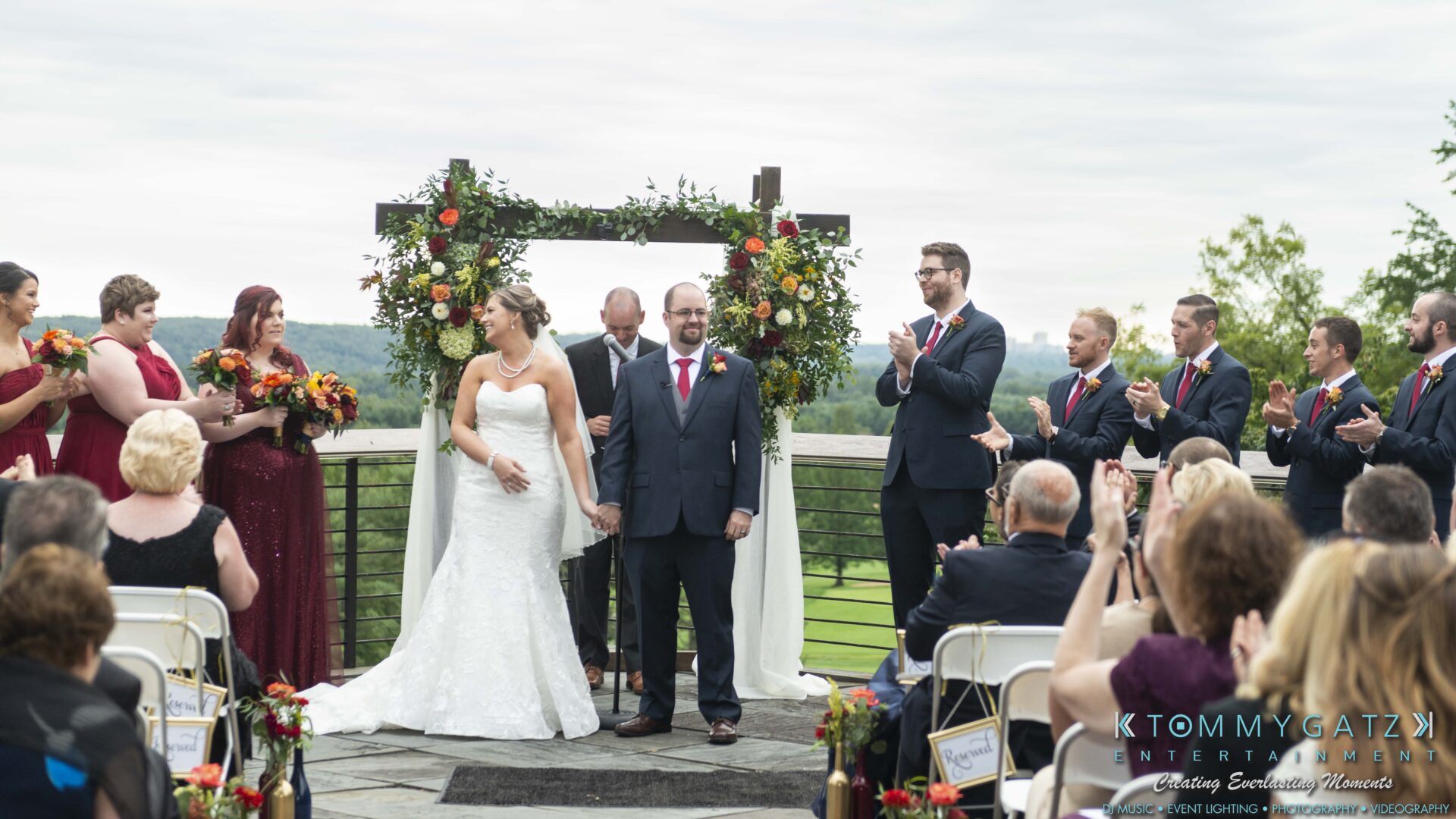 Lisa-Robert-092218-BLOG-selections-WM-59 - TOMMY GATZ ENTERTAINMENT A bride and groom stand together at an outdoor wedding ceremony, surrounded by a bridal party and guests applauding. A floral arch and scenic landscape are in the background.