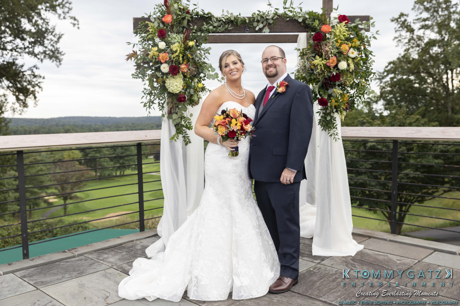 Lisa-Robert-092218-BLOG-selections-WM-61 - TOMMY GATZ ENTERTAINMENT A couple poses in wedding attire under a floral arch on a terrace, with a scenic view in the background.