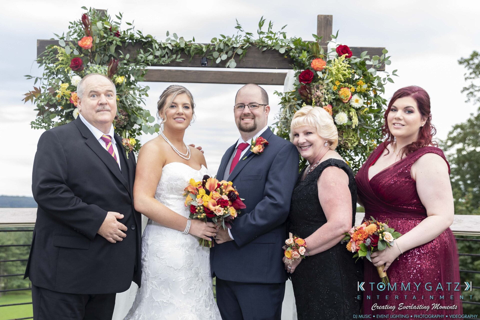 Lisa-Robert-092218-BLOG-selections-WM-62 - TOMMY GATZ ENTERTAINMENT A wedding group photo with two couples and one woman in formal attire, standing in front of a floral archway outdoors.