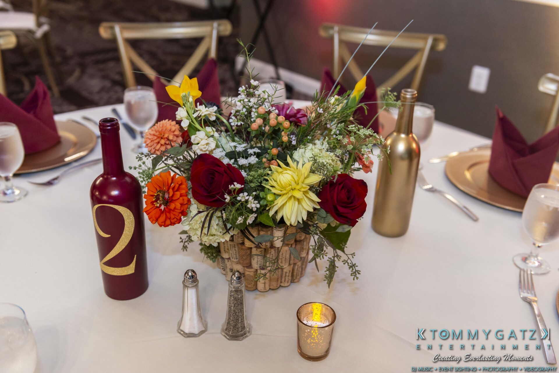 Lisa-Robert-092218-BLOG-selections-WM-65 - TOMMY GATZ ENTERTAINMENT Table centerpiece with colorful flowers in a vase made of wine corks. Two wine bottles, one red with the number two, and gold one. Set dining table with folded napkins and utensils.