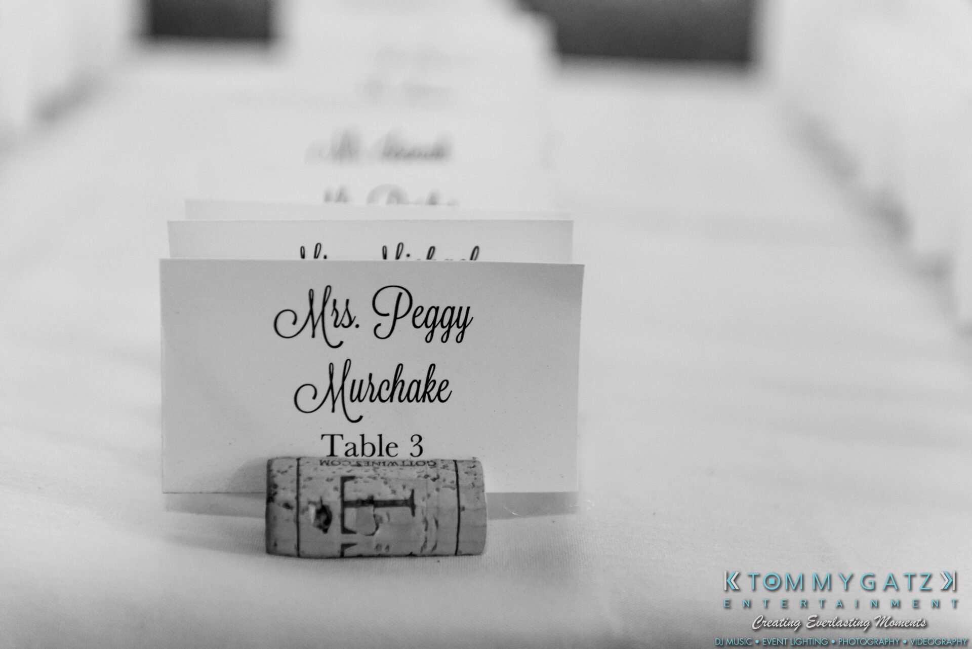 Lisa-Robert-092218-BLOG-selections-WM-66 - TOMMY GATZ ENTERTAINMENT Place card on a table displays the name "Mrs. Peggy Murchake" and "Table 3," held by a wine cork. Tommy Gatz Entertainment watermark visible in the bottom right corner.