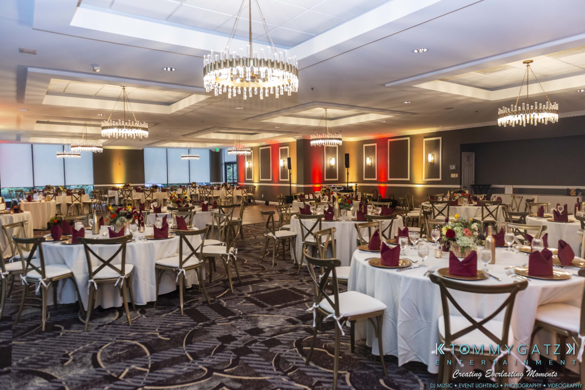 Lisa-Robert-092218-BLOG-selections-WM-67 - TOMMY GATZ ENTERTAINMENT A banquet hall with round tables set for an event, featuring white tablecloths, burgundy napkins, and wooden chairs. Chandeliers hang from the ceiling, providing elegant lighting.
