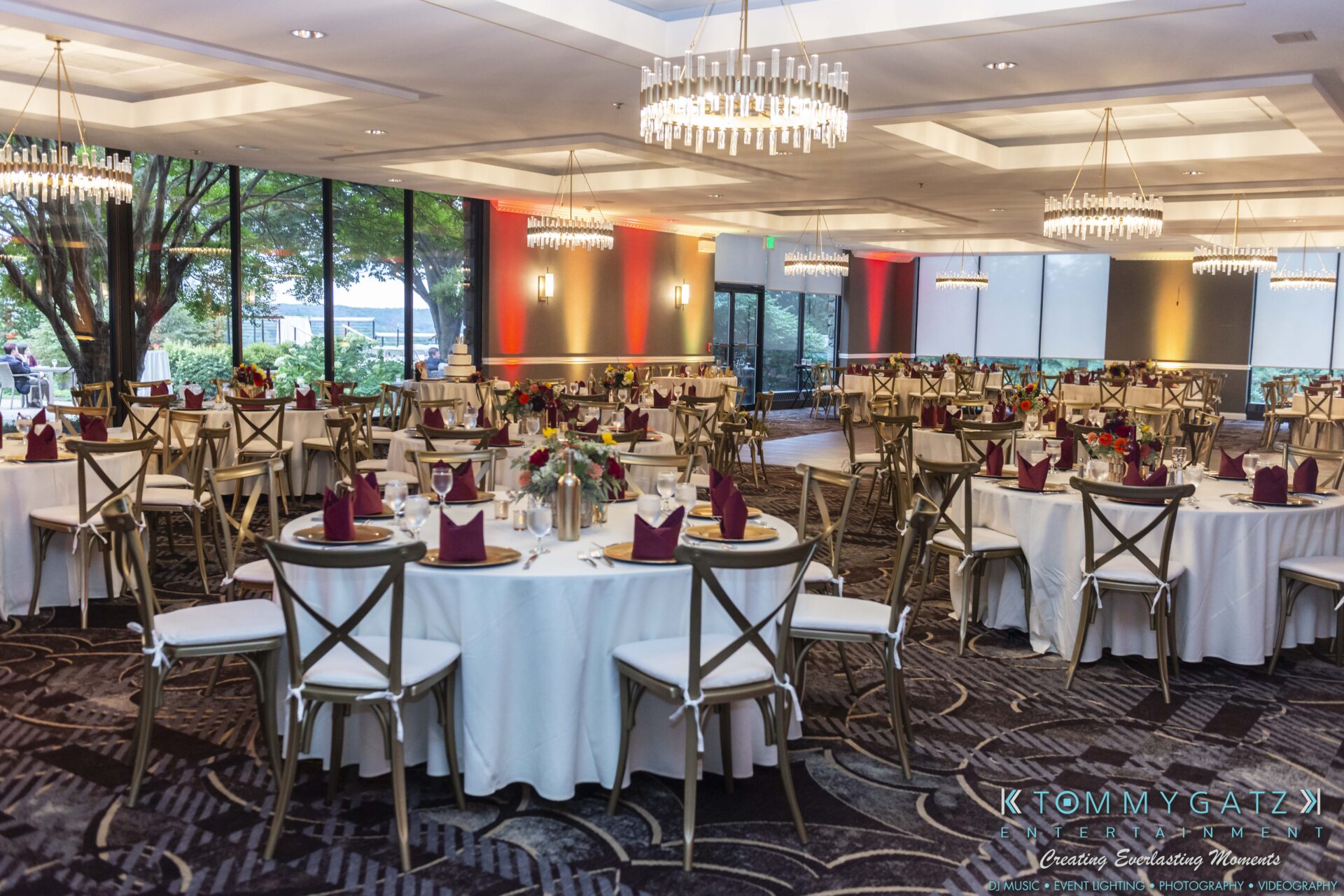 Lisa-Robert-092218-BLOG-selections-WM-68 - TOMMY GATZ ENTERTAINMENT Elegant banquet hall with round tables set for an event, featuring white tablecloths, burgundy napkins, and floral centerpieces, illuminated by chandeliers and natural light from large windows.