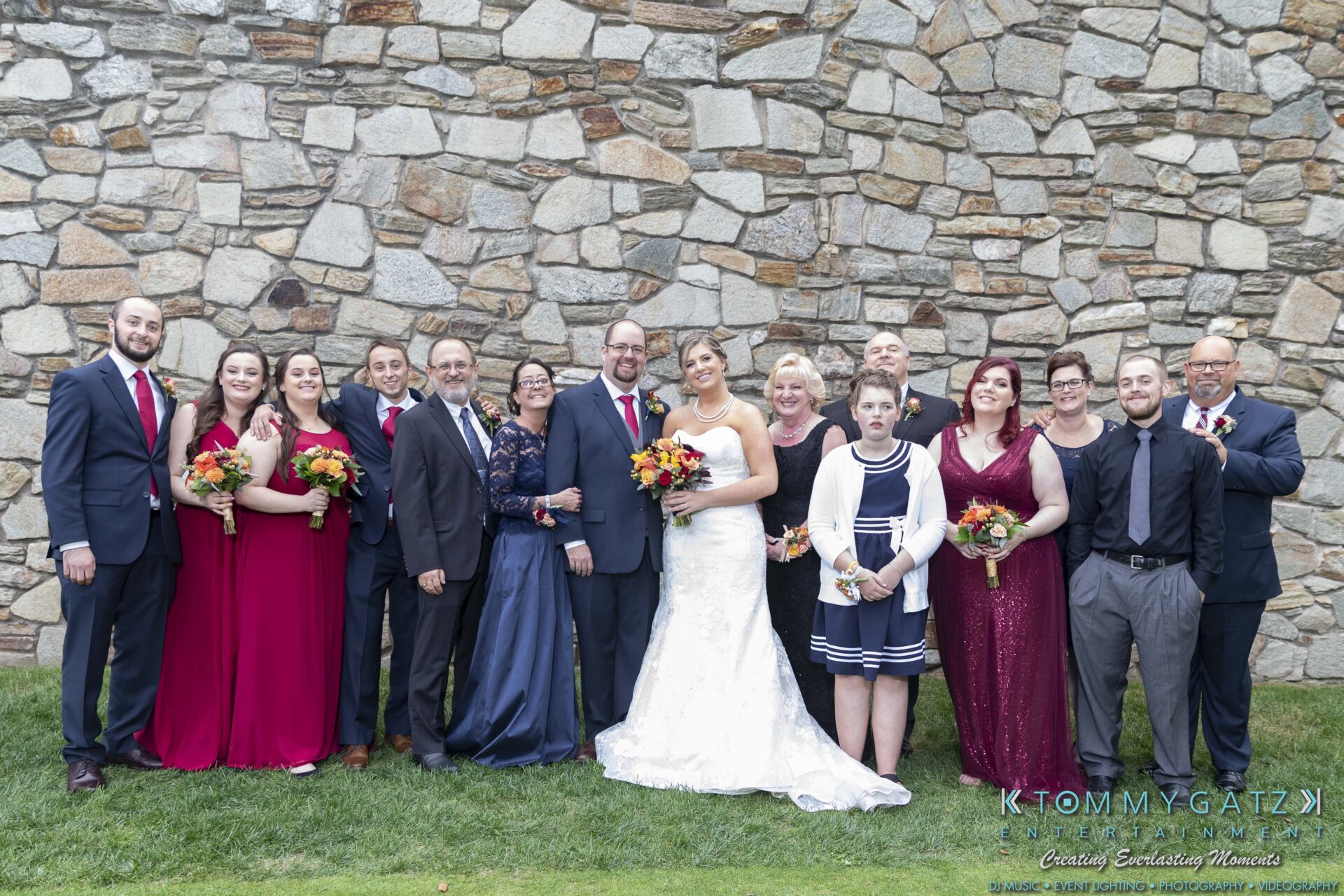 Lisa-Robert-092218-BLOG-selections-WM-69 - TOMMY GATZ ENTERTAINMENT A group of people, including a bride and groom, pose in formal attire outdoors in front of a stone wall. Some hold bouquets.