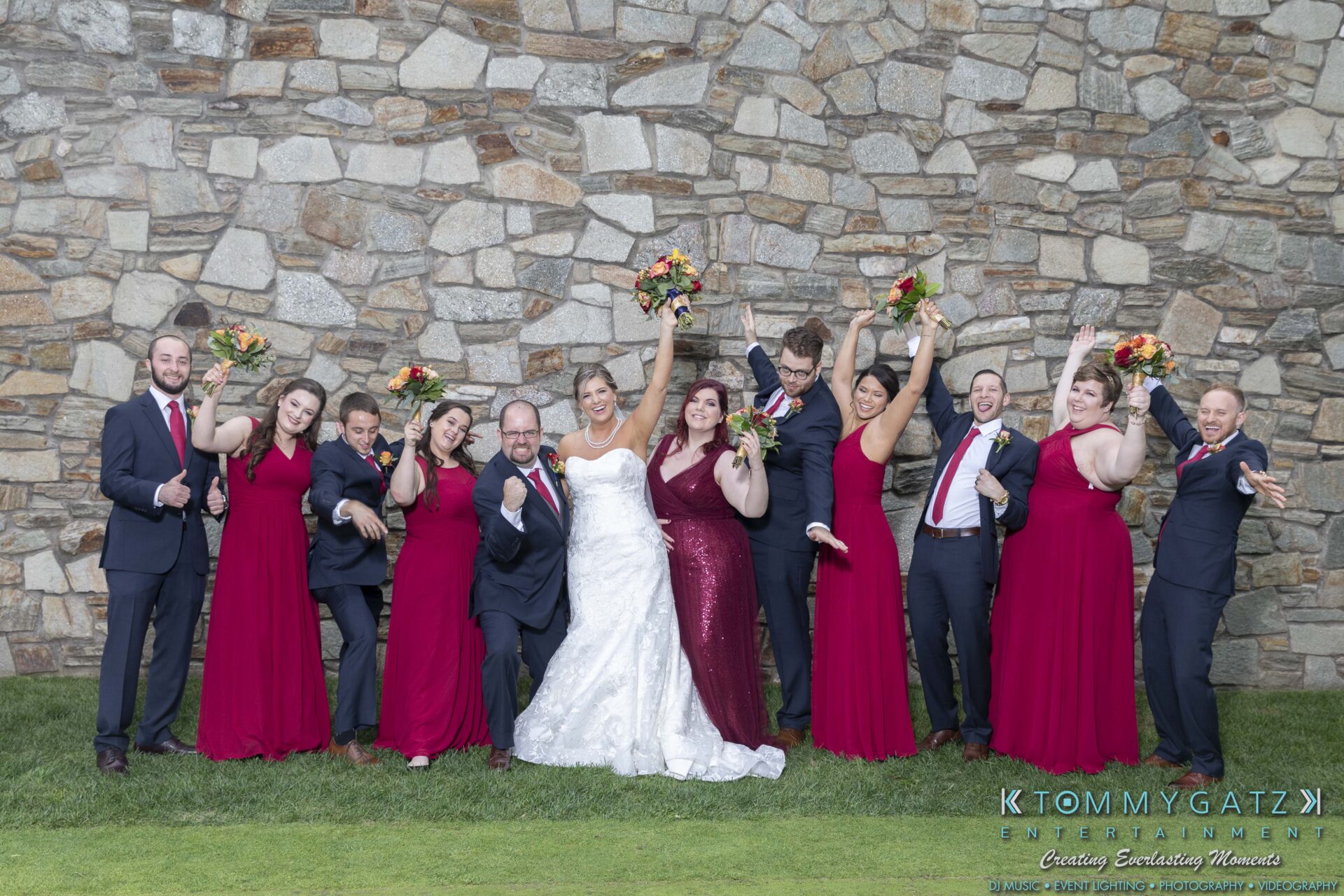 Lisa-Robert-092218-BLOG-selections-WM-71 - TOMMY GATZ ENTERTAINMENT A wedding party poses outdoors against a stone wall. The bride and groom are in the center, surrounded by bridesmaids in burgundy dresses and groomsmen in black suits holding bouquets.