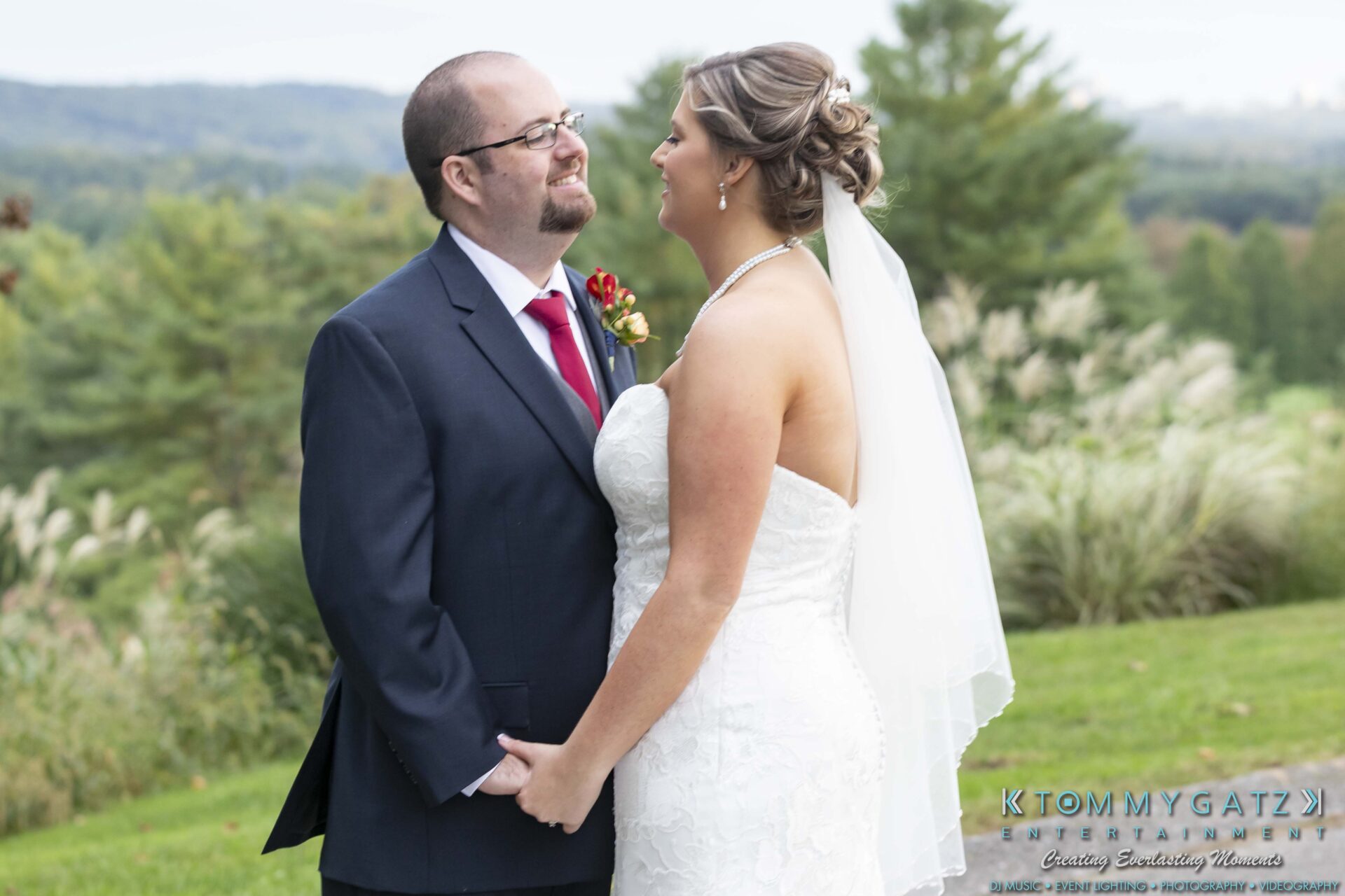 Lisa-Robert-092218-BLOG-selections-WM-74 - TOMMY GATZ ENTERTAINMENT A bride and groom stand outdoors, holding hands and smiling at each other, with a scenic background of greenery and distant hills.