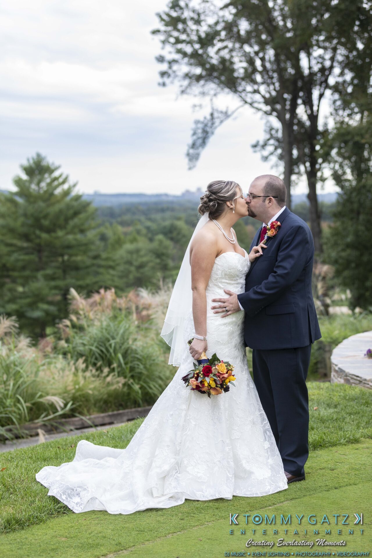 Lisa-Robert-092218-BLOG-selections-WM-76 - TOMMY GATZ ENTERTAINMENT A bride and groom kissing outdoors in a garden setting. The bride holds a bouquet of colorful flowers, and the groom is in a dark suit. Trees and landscape are visible in the background.