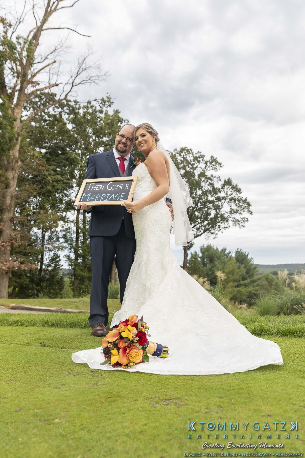 Lisa-Robert-092218-BLOG-selections-WM-78 - TOMMY GATZ ENTERTAINMENT Bride and groom stand outdoors holding a sign reading, "Then comes marriage," with a bouquet on the grass in front of them.