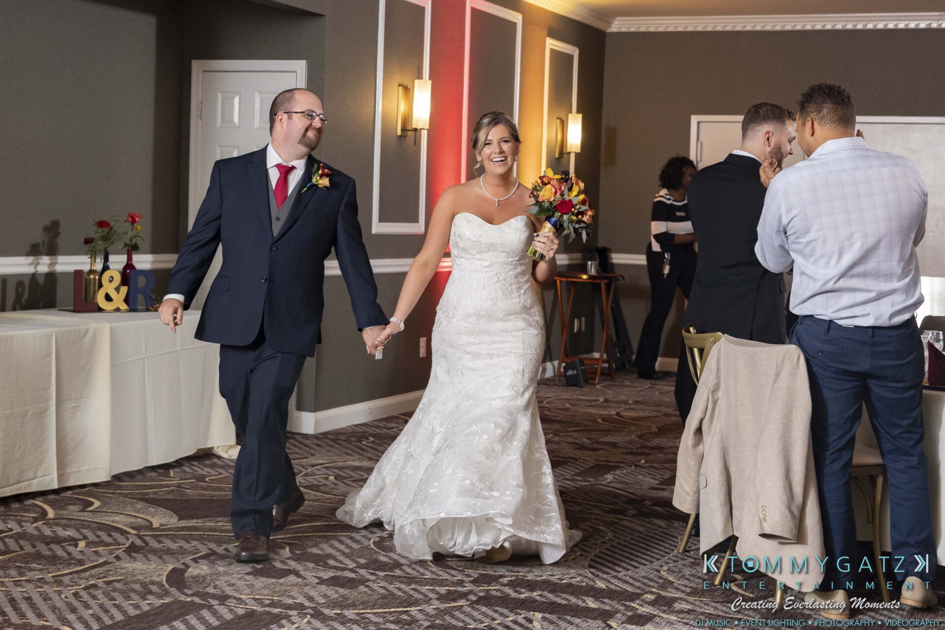 Lisa-Robert-092218-BLOG-selections-WM-83 - TOMMY GATZ ENTERTAINMENT A bride and groom enter a decorated room, smiling and holding hands. The bride wears a white gown, and the groom is in a suit. Guests are visible in the background.