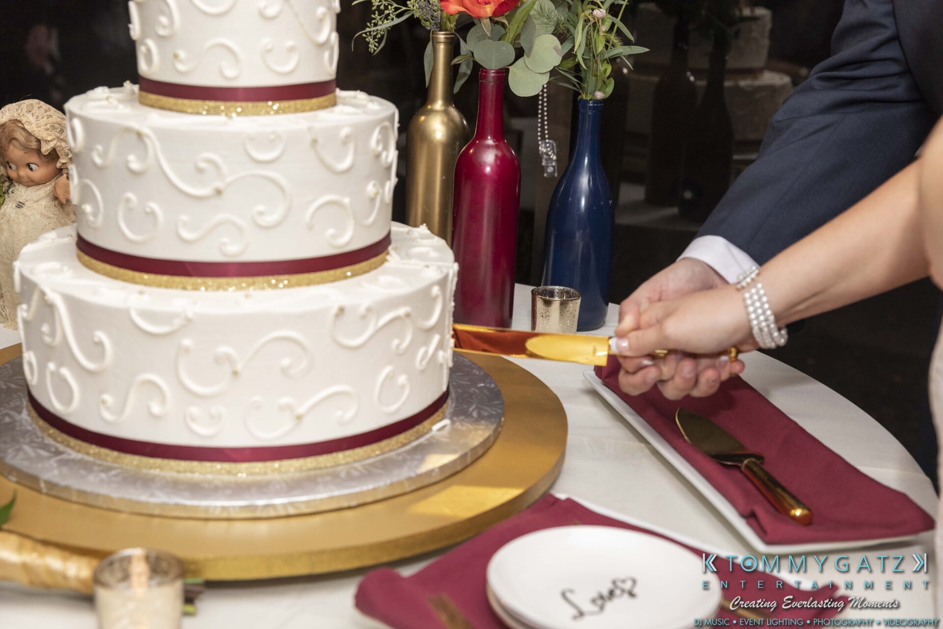 Lisa-Robert-092218-BLOG-selections-WM-94 - TOMMY GATZ ENTERTAINMENT A couple cuts a three-tiered white cake with a gold knife. The table is set with plates and red napkins.