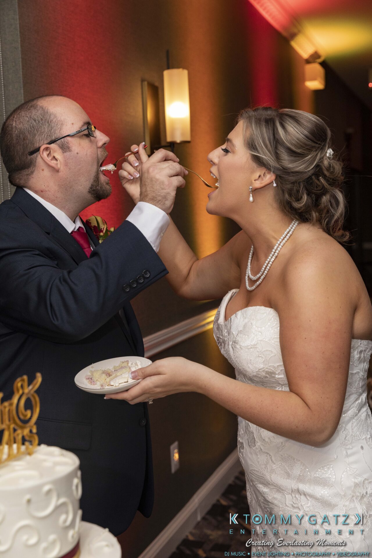 Lisa-Robert-092218-BLOG-selections-WM-96 - TOMMY GATZ ENTERTAINMENT A bride and groom feed each other cake at a wedding, standing next to a decorated table.