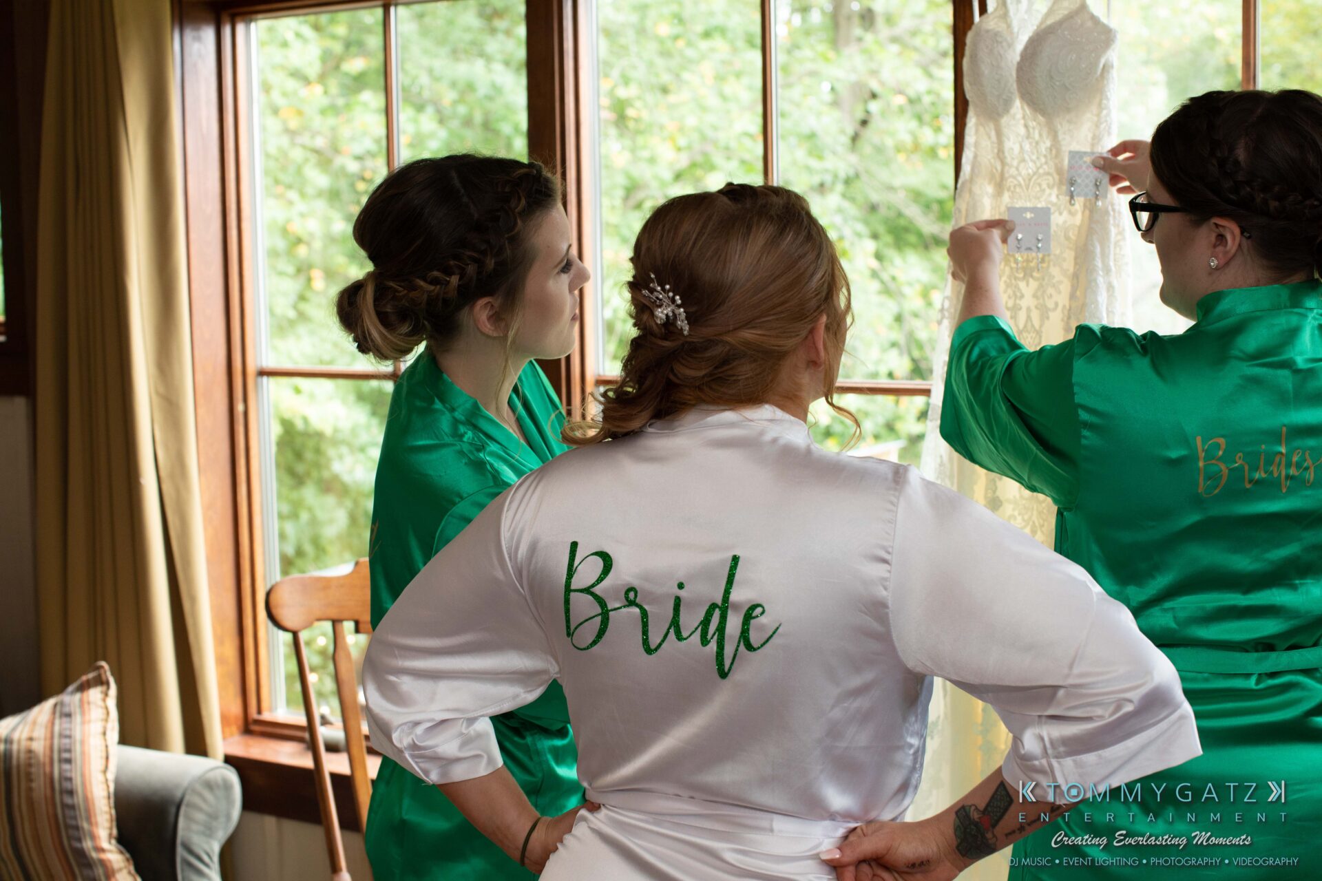 Proctor-Wedding-HIGHLIGHTS-091419-1 - TOMMY GATZ ENTERTAINMENT Three women in green and white robes labeled "Bride" look at a hanging white lace dress in a room with large windows and wooden trim.