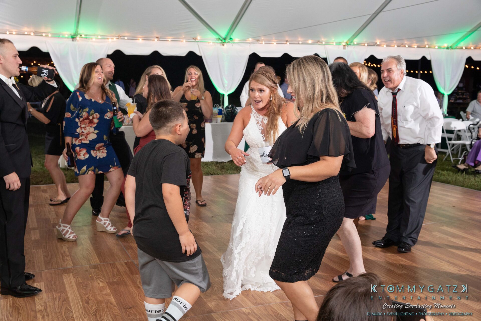 Proctor-Wedding-HIGHLIGHTS-091419-102 - TOMMY GATZ ENTERTAINMENT People dancing enthusiastically on a wooden dance floor under a white tent with string lights at a wedding reception.