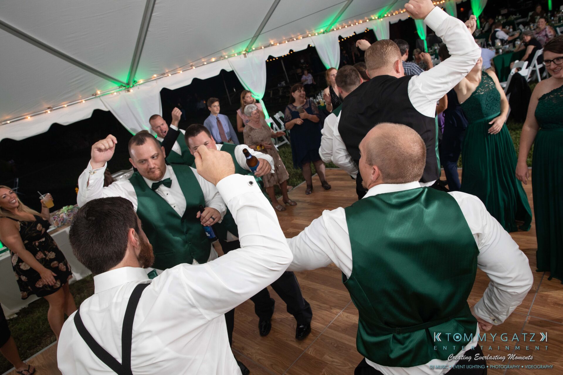 Proctor-Wedding-HIGHLIGHTS-091419-103 - TOMMY GATZ ENTERTAINMENT Guests in green vests energetically dance at a formal event under a white tent, with other attendees and string lights in the background.