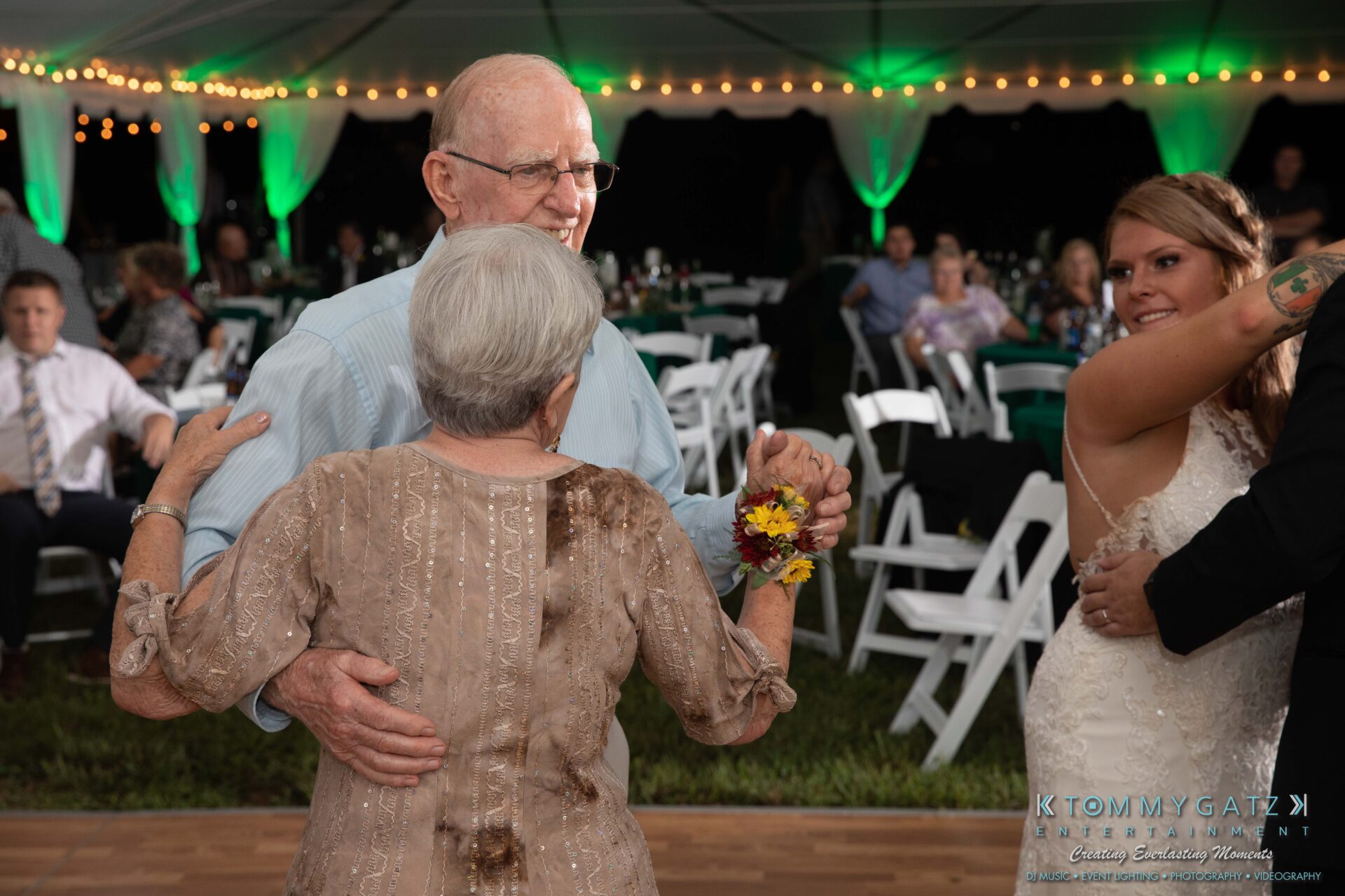 Proctor-Wedding-HIGHLIGHTS-091419-105 - TOMMY GATZ ENTERTAINMENT An elderly couple dances under a tent at an outdoor event, surrounded by tables and chairs. A bride and another person are dancing nearby.