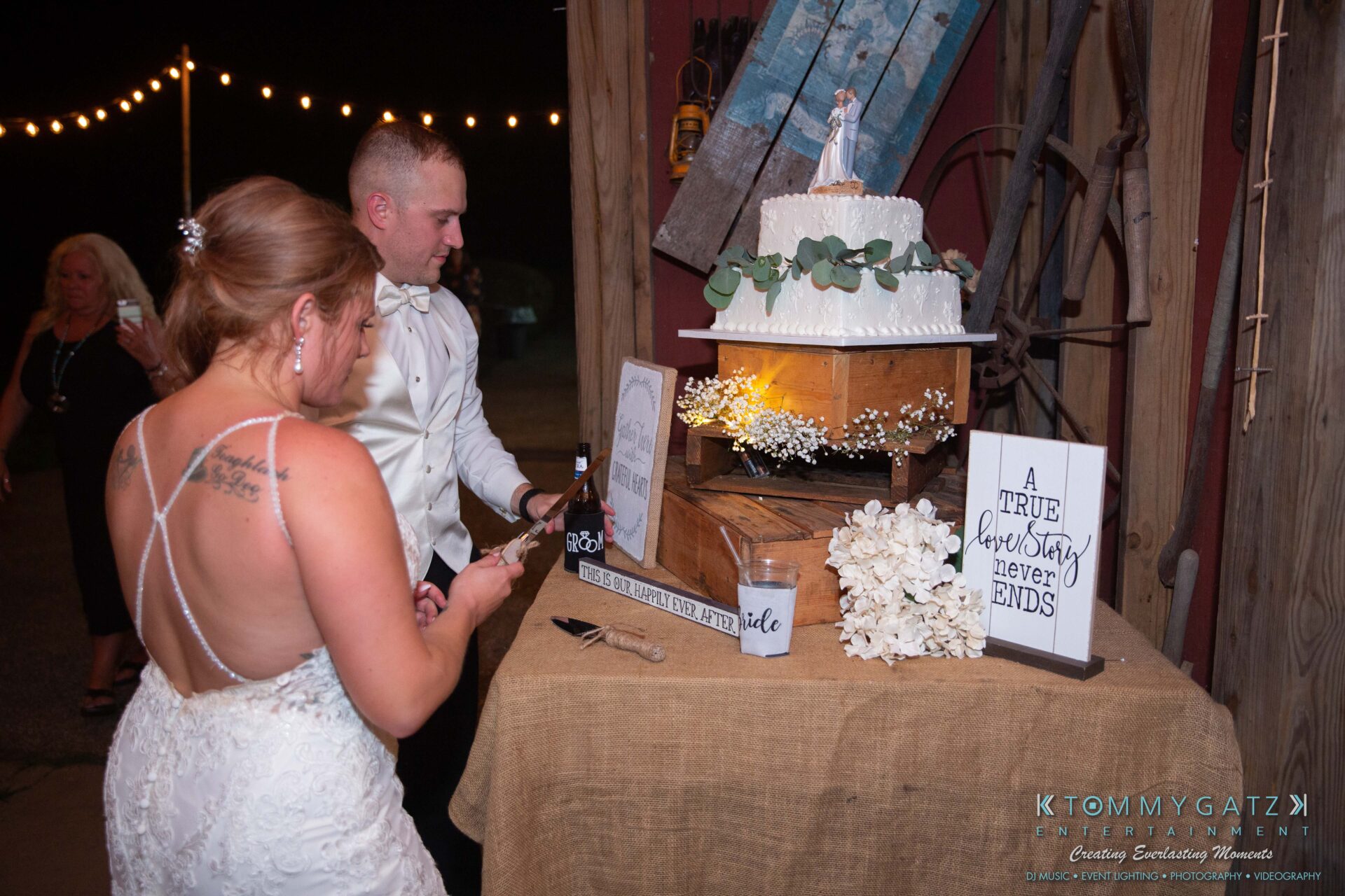 Proctor-Wedding-HIGHLIGHTS-091419-110 - TOMMY GATZ ENTERTAINMENT A bride and groom prepare to cut a tiered wedding cake adorned with flowers. A sign reads "A true love story never ends." String lights and a wooden backdrop are in the background.