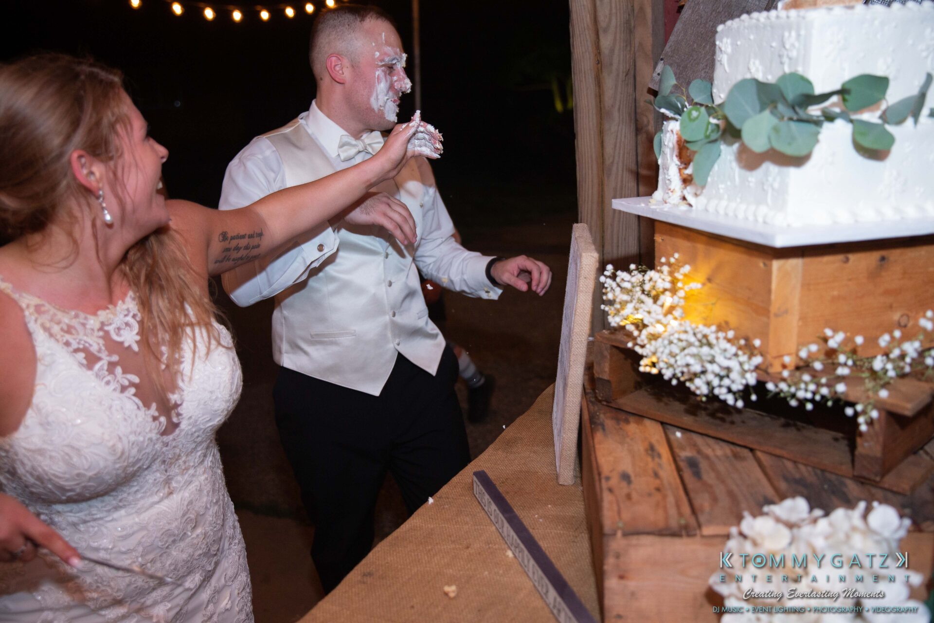 Proctor-Wedding-HIGHLIGHTS-091419-111 - TOMMY GATZ ENTERTAINMENT Bride playfully smears cake on groom's face at a wedding reception. Cake is placed on a rustic wooden stand decorated with greenery and baby's breath flowers.