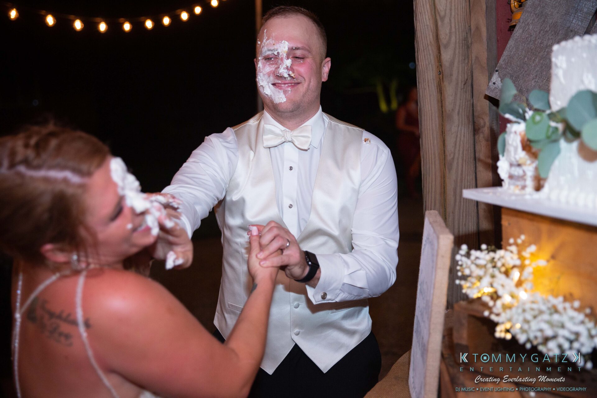 Proctor-Wedding-HIGHLIGHTS-091419-112 - TOMMY GATZ ENTERTAINMENT A bride playfully smears cake on the groom's face during a wedding reception. They are both smiling, with cake on a table nearby.