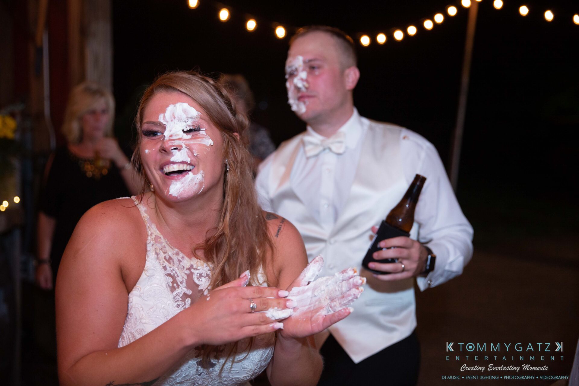 Proctor-Wedding-HIGHLIGHTS-091419-113 - TOMMY GATZ ENTERTAINMENT A couple with cake smeared on their faces smiles at an outdoor event. The woman laughs with her hands covered in cake, while the man holds a drink. String lights hang above.