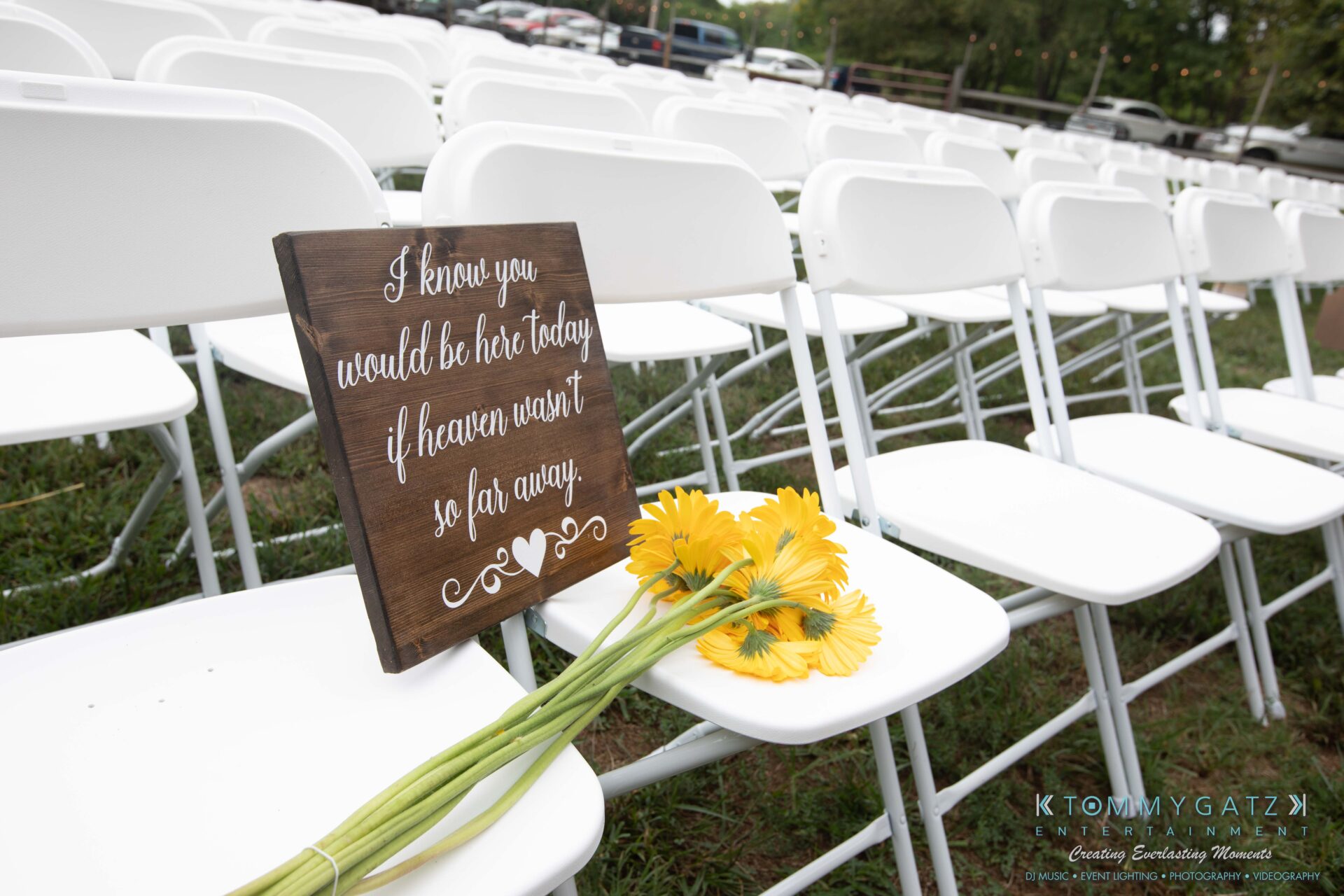 Proctor-Wedding-HIGHLIGHTS-091419-12 - TOMMY GATZ ENTERTAINMENT Close-up of white folding chairs with a wooden sign reading, "I know you would be here today if heaven wasn’t so far away," and a bouquet of yellow flowers resting on a seat.