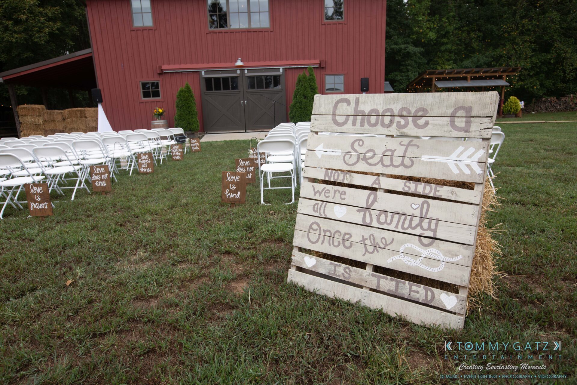 Proctor-Wedding-HIGHLIGHTS-091419-13 - TOMMY GATZ ENTERTAINMENT Outdoor wedding setup with white chairs facing a red barn. A rustic wooden sign reads, "Choose a seat, not a side, we are family once the knot is tied.