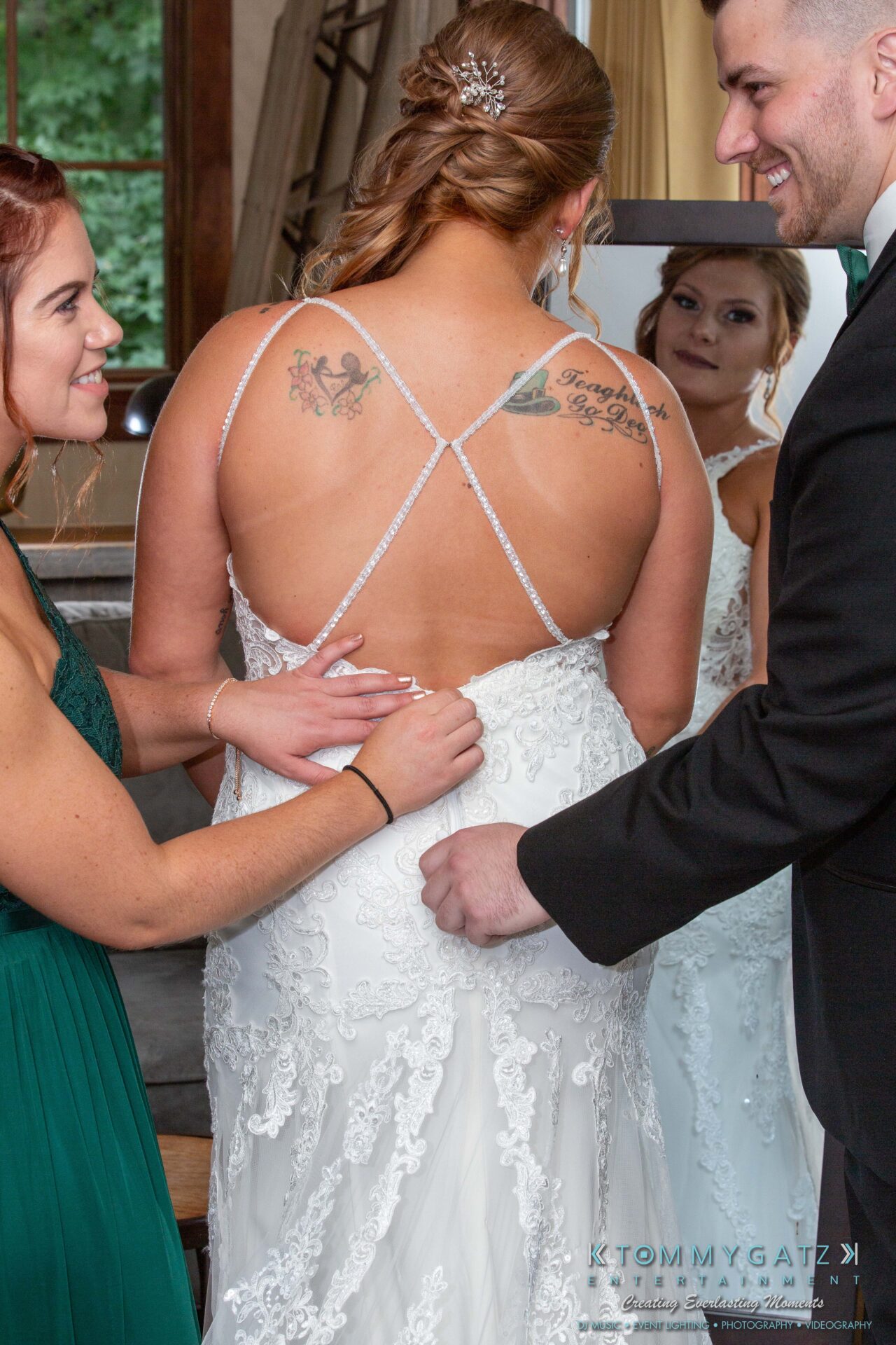 Proctor-Wedding-HIGHLIGHTS-091419-17 - TOMMY GATZ ENTERTAINMENT Bride in white dress with floral back tattoos, assisted by a woman in green dress and a man in a suit. They are smiling in front of a mirror.