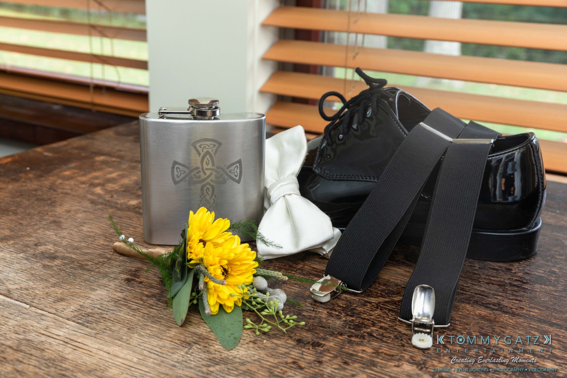 Proctor-Wedding-HIGHLIGHTS-091419-21 - TOMMY GATZ ENTERTAINMENT Grooming accessories on a wooden table: black shoes, suspenders, a white bow tie, a metal flask with an engraved design, and a yellow flower boutonniere.