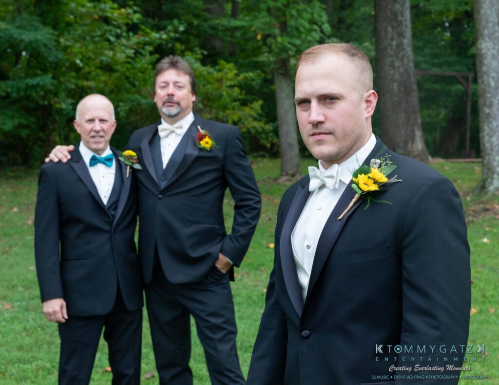 Proctor-Wedding-HIGHLIGHTS-091419-31 - TOMMY GATZ ENTERTAINMENT Three men in tuxedos with floral boutonnieres stand on a grassy area surrounded by trees. One man is in the foreground, and two are in the background.