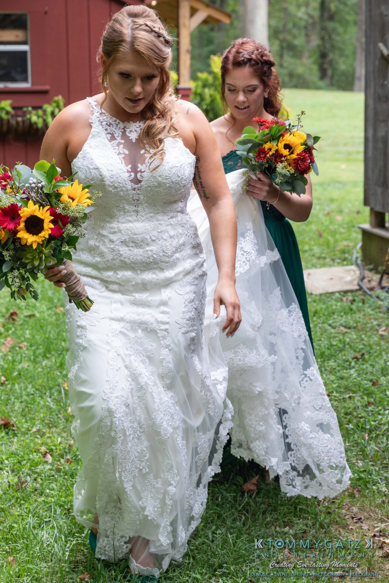 Proctor-Wedding-HIGHLIGHTS-091419-32 1 - TOMMY GATZ ENTERTAINMENT A bride in a white lace gown holds a bouquet with sunflowers, walking alongside a bridesmaid in a green dress, also holding a bouquet, on a grassy outdoor setting.