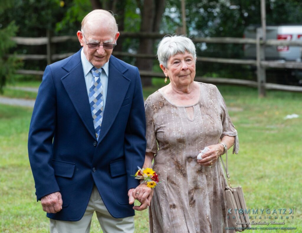 Proctor-Wedding-HIGHLIGHTS-091419-36 - TOMMY GATZ ENTERTAINMENT An elderly couple walks hand in hand outdoors; the man wears a blue blazer and tie, and the woman wears a light-colored dress, holding a small bouquet.