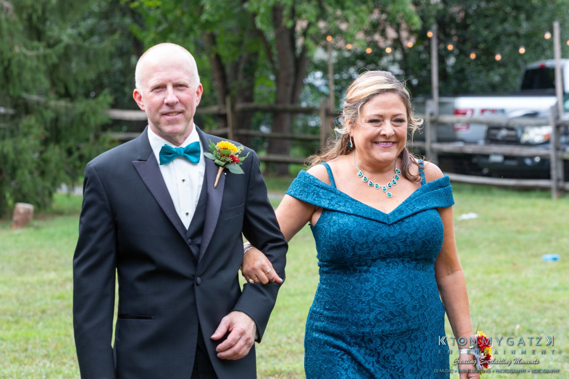 Proctor-Wedding-HIGHLIGHTS-091419-37 - TOMMY GATZ ENTERTAINMENT A man in a tuxedo and a woman in a teal dress walk arm in arm outdoors, with trees and a wooden fence in the background.