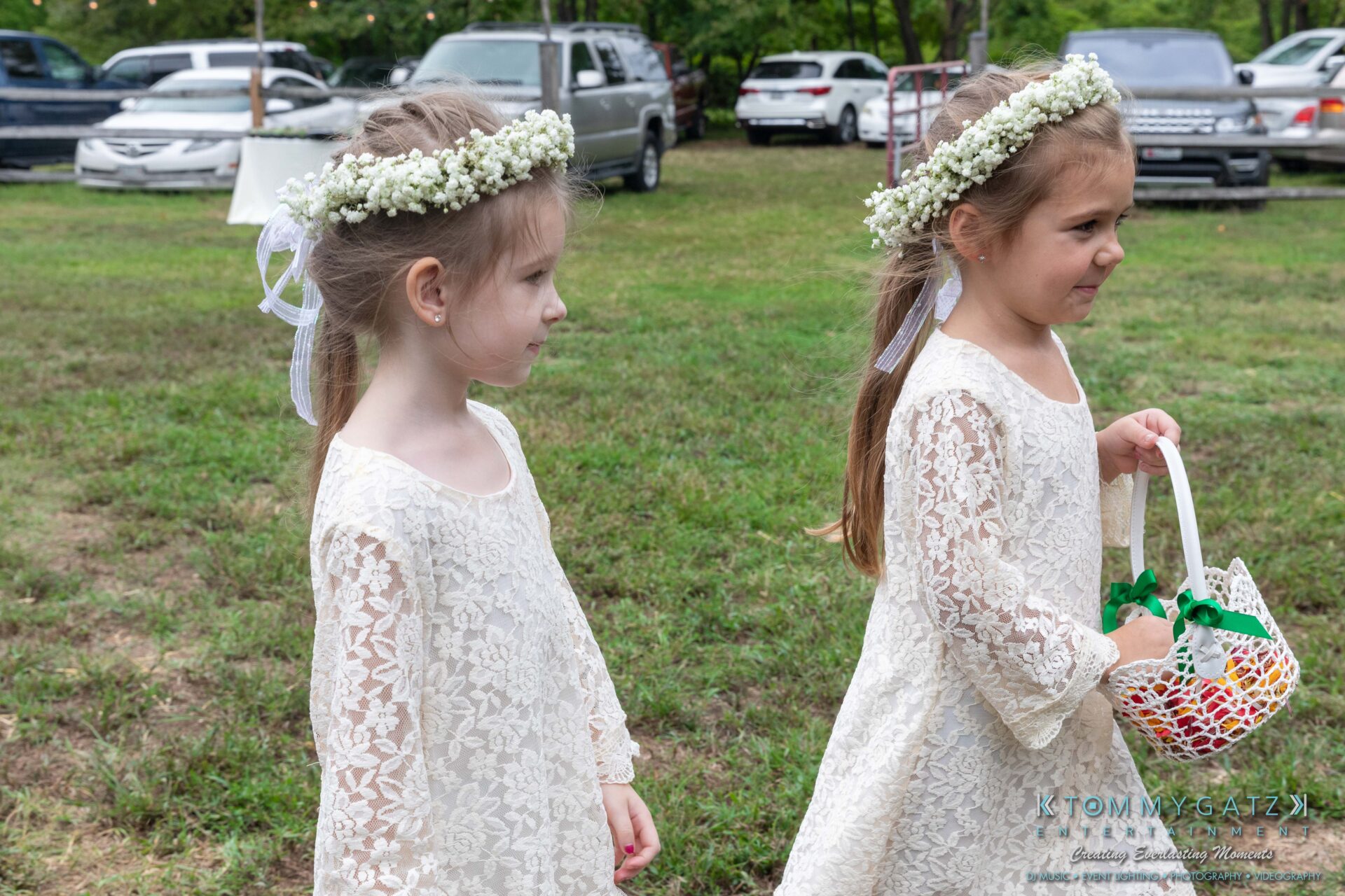 Proctor-Wedding-HIGHLIGHTS-091419-42 - TOMMY GATZ ENTERTAINMENT Two young girls in white lace dresses and flower crowns walk on grass. One holds a white basket with green ribbons. Cars and trees are in the background.