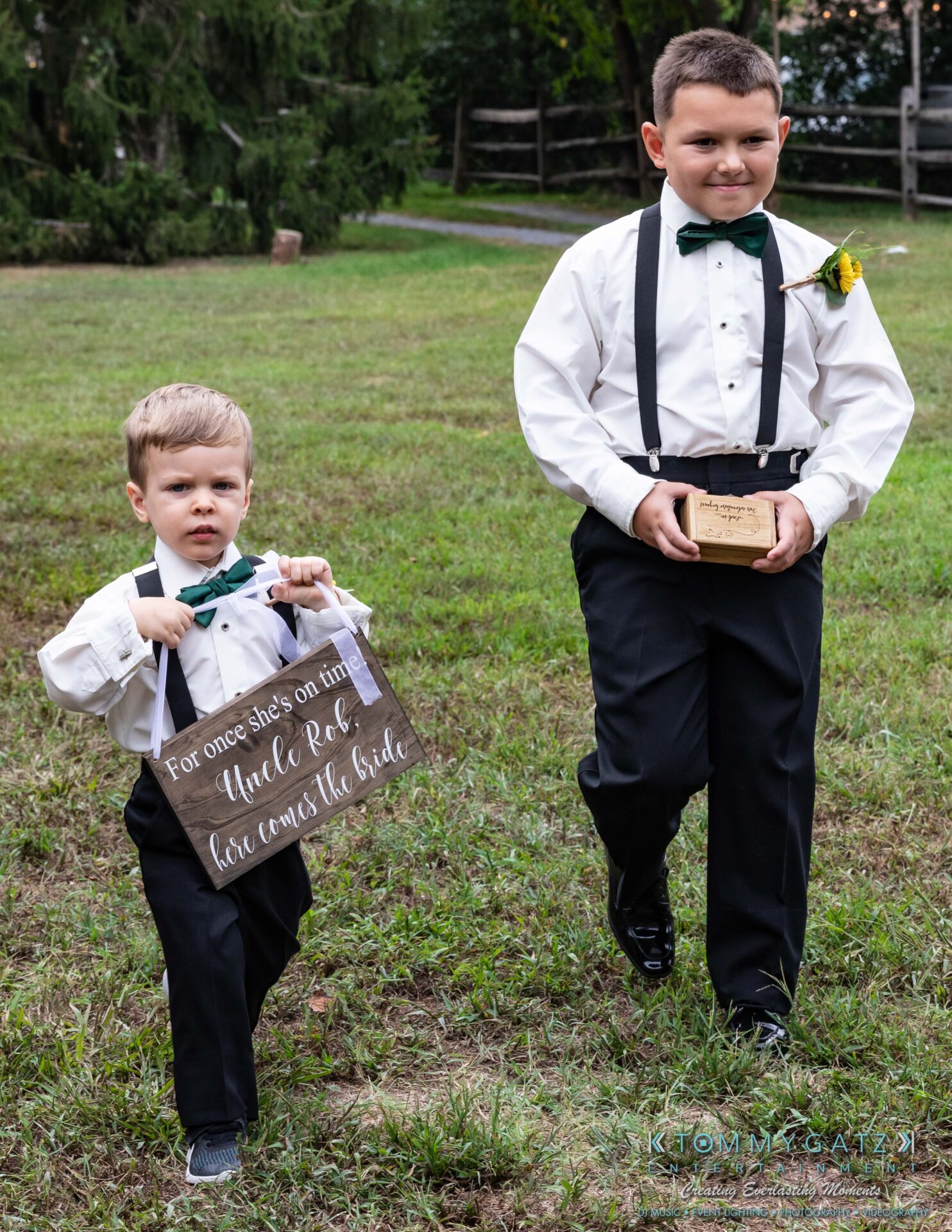 Proctor-Wedding-HIGHLIGHTS-091419-43 - TOMMY GATZ ENTERTAINMENT Two young boys dressed in formal attire walk on grass. One holds a sign saying "For once she's on time, Uncle Rob, here comes the bride." The other carries a wooden box.