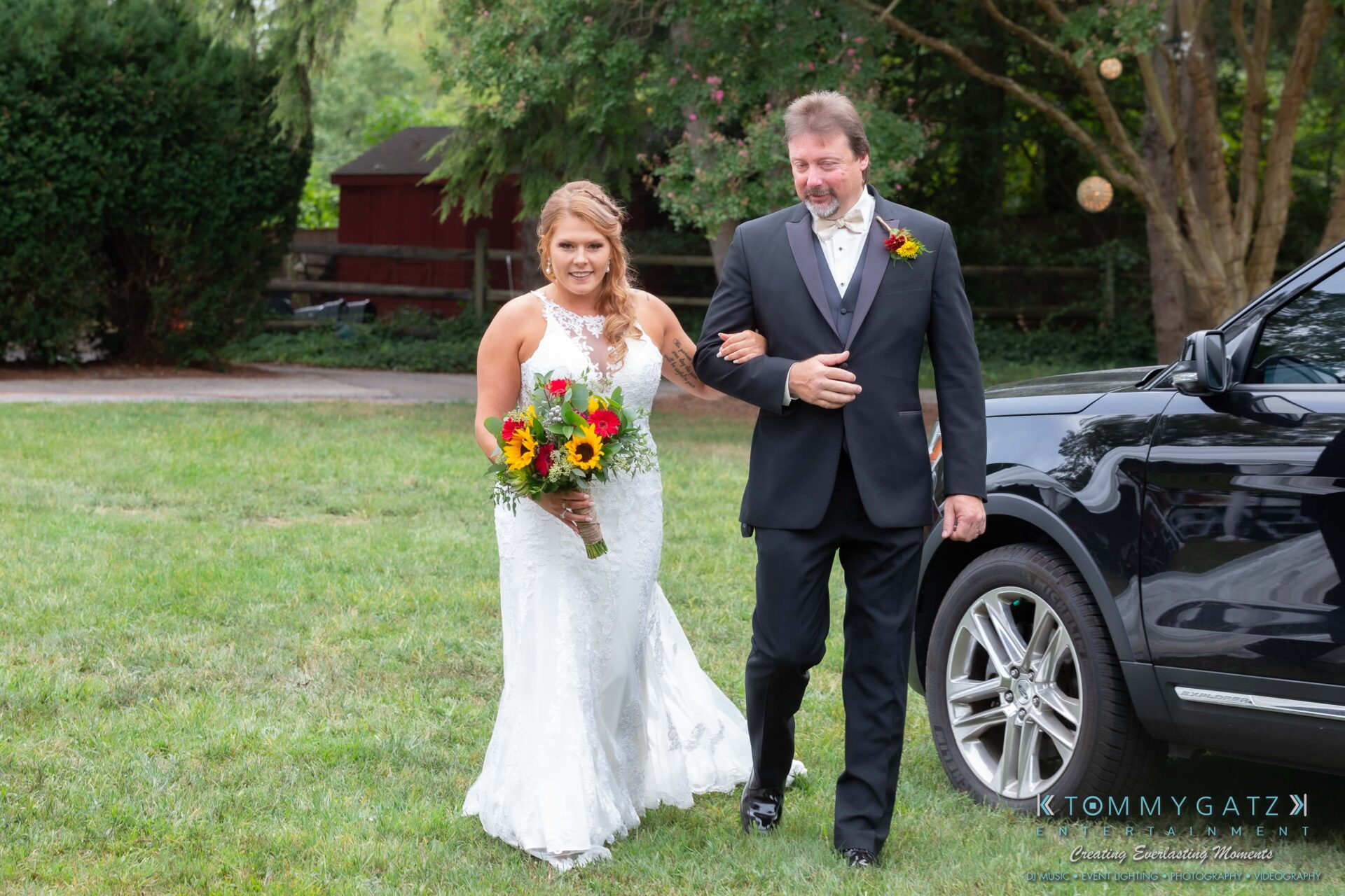 Proctor-Wedding-HIGHLIGHTS-091419-45 - TOMMY GATZ ENTERTAINMENT A bride in a white dress, holding a bouquet, walks arm-in-arm with a man in a suit next to a black car on a grassy area.