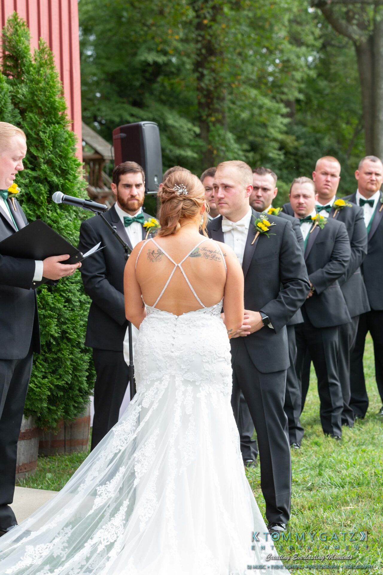 Proctor-Wedding-HIGHLIGHTS-091419-50 - TOMMY GATZ ENTERTAINMENT A bride and groom stand facing each other during an outdoor wedding ceremony, with the officiant and groomsmen in the background.