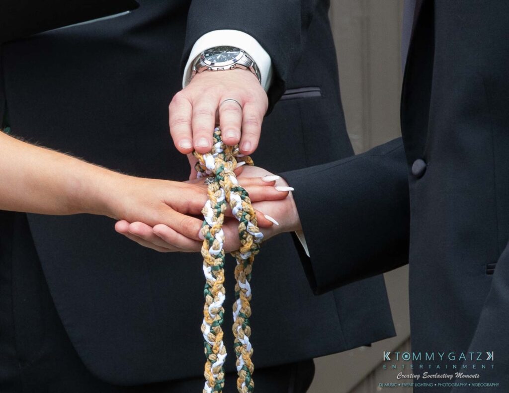 Proctor-Wedding-HIGHLIGHTS-091419-55 - TOMMY GATZ ENTERTAINMENT Three hands hold intertwined cords, symbolizing unity, in a formal setting.