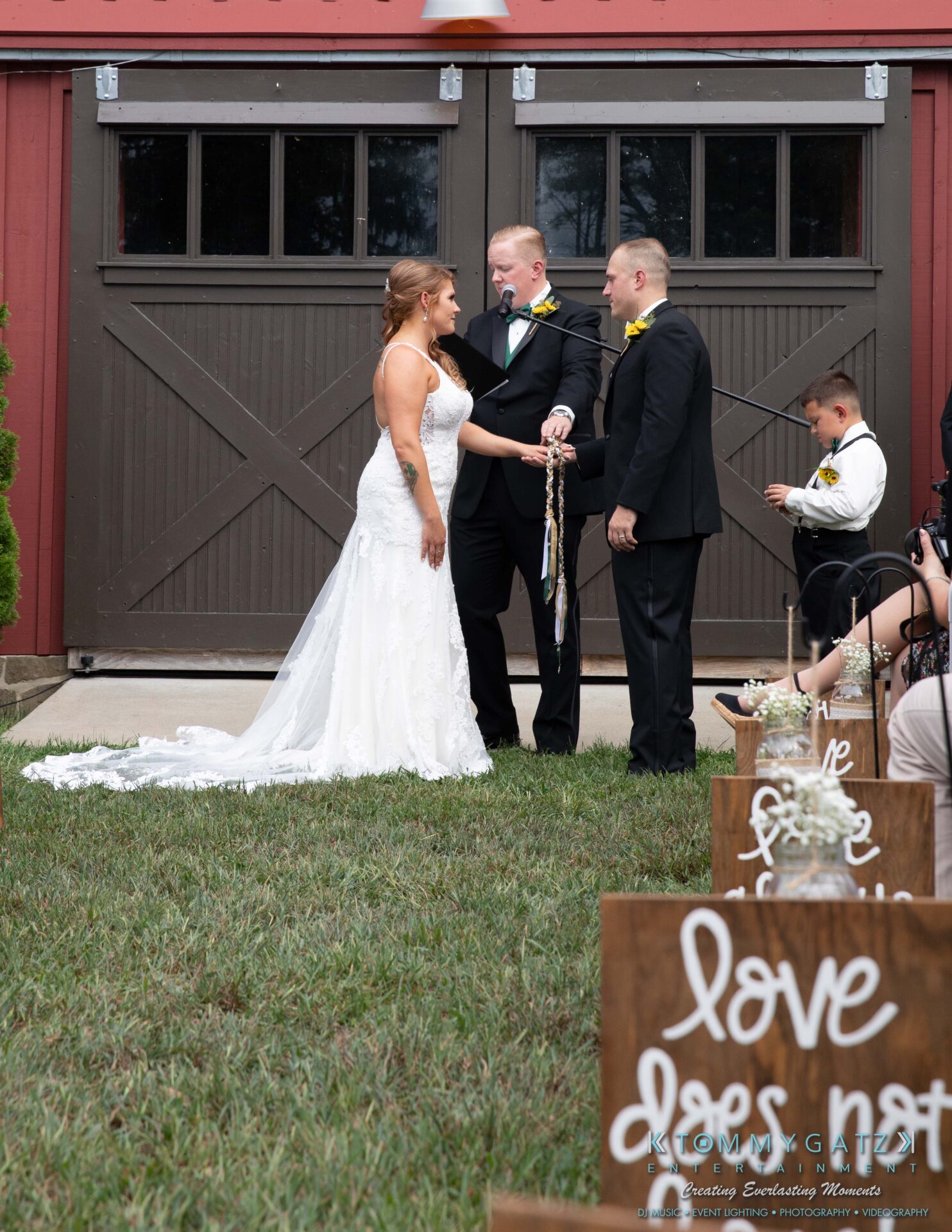 Proctor-Wedding-HIGHLIGHTS-091419-56 - TOMMY GATZ ENTERTAINMENT A couple exchanges vows in an outdoor wedding ceremony in front of a barn. A young boy sits nearby, and wooden signs with text are placed on the grass.
