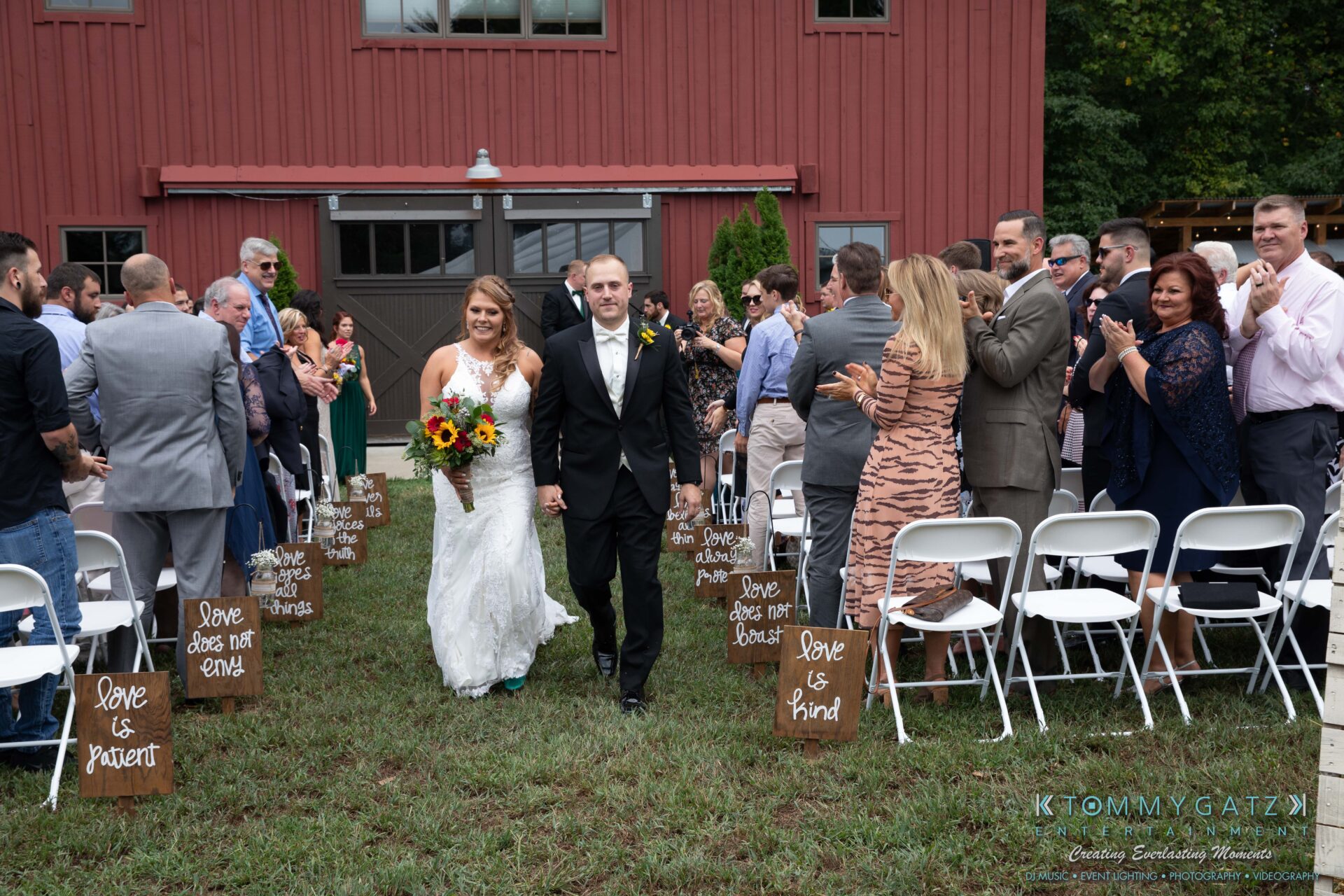 Proctor-Wedding-HIGHLIGHTS-091419-57 - TOMMY GATZ ENTERTAINMENT A bride and groom walk down the aisle outdoors. Guests, seated in white chairs, clap and smile. Wooden barn-like building in the background.