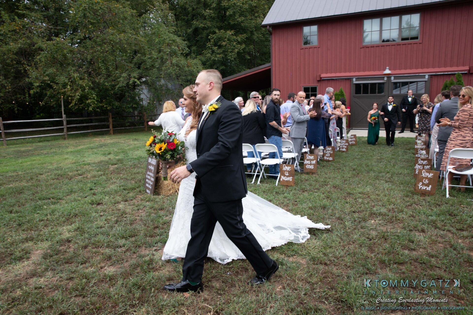 Proctor-Wedding-HIGHLIGHTS-091419-58 - TOMMY GATZ ENTERTAINMENT A bride and groom walk hand in hand down the aisle outdoors, with guests seated on either side. A barn is visible in the background.