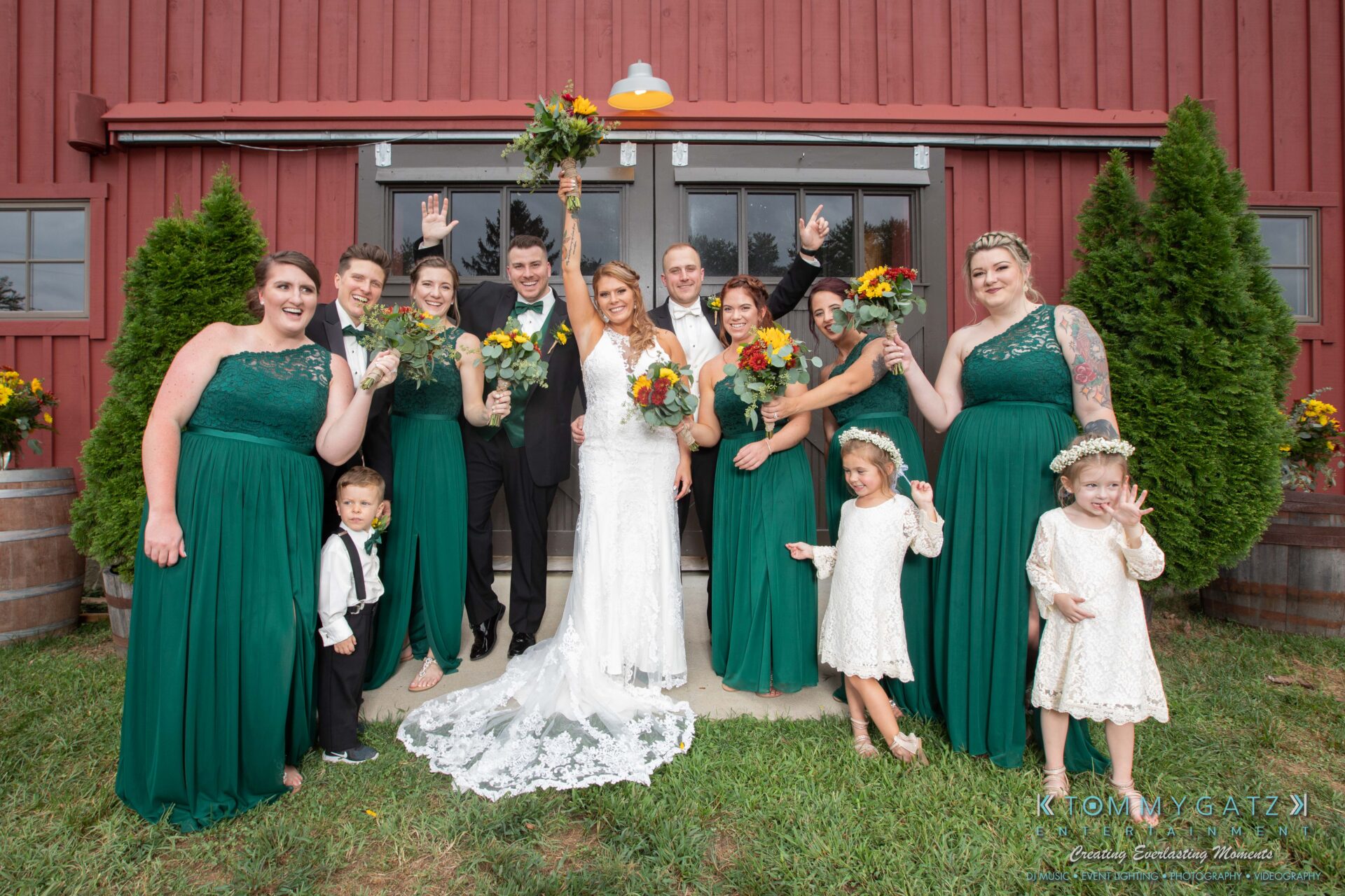 Proctor-Wedding-HIGHLIGHTS-091419-65 - TOMMY GATZ ENTERTAINMENT A wedding party poses in front of a red barn. The bride and groom, surrounded by bridesmaids in green dresses and groomsmen in dark suits, are smiling and holding bouquets.
