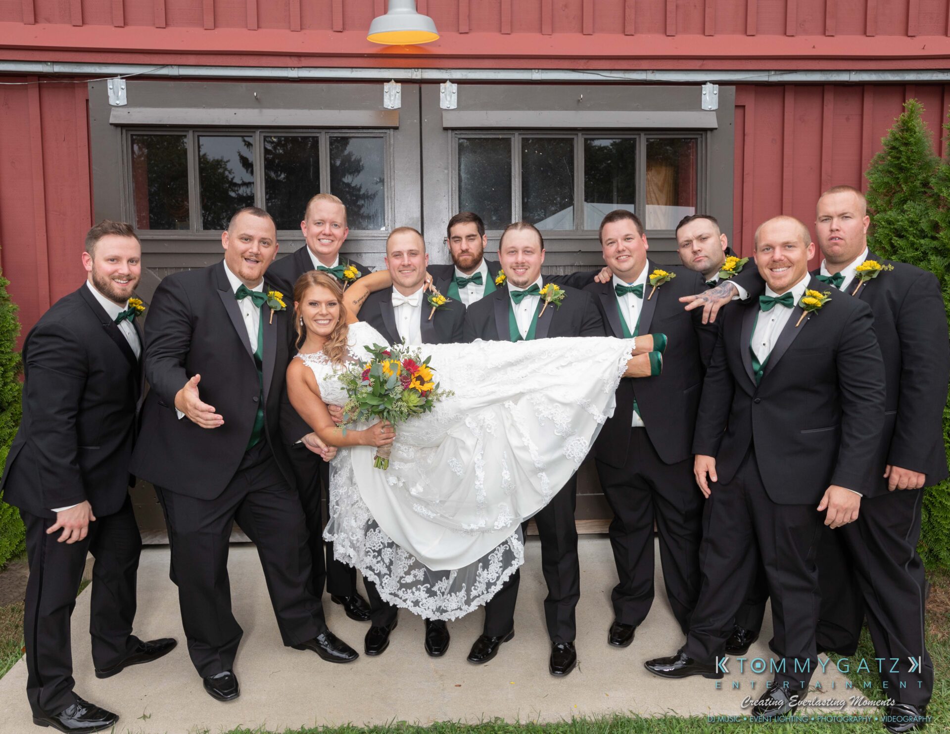 Proctor-Wedding-HIGHLIGHTS-091419-66 - TOMMY GATZ ENTERTAINMENT A bride in a white dress is playfully lifted by groomsmen in black suits and green vests in front of a rustic building.