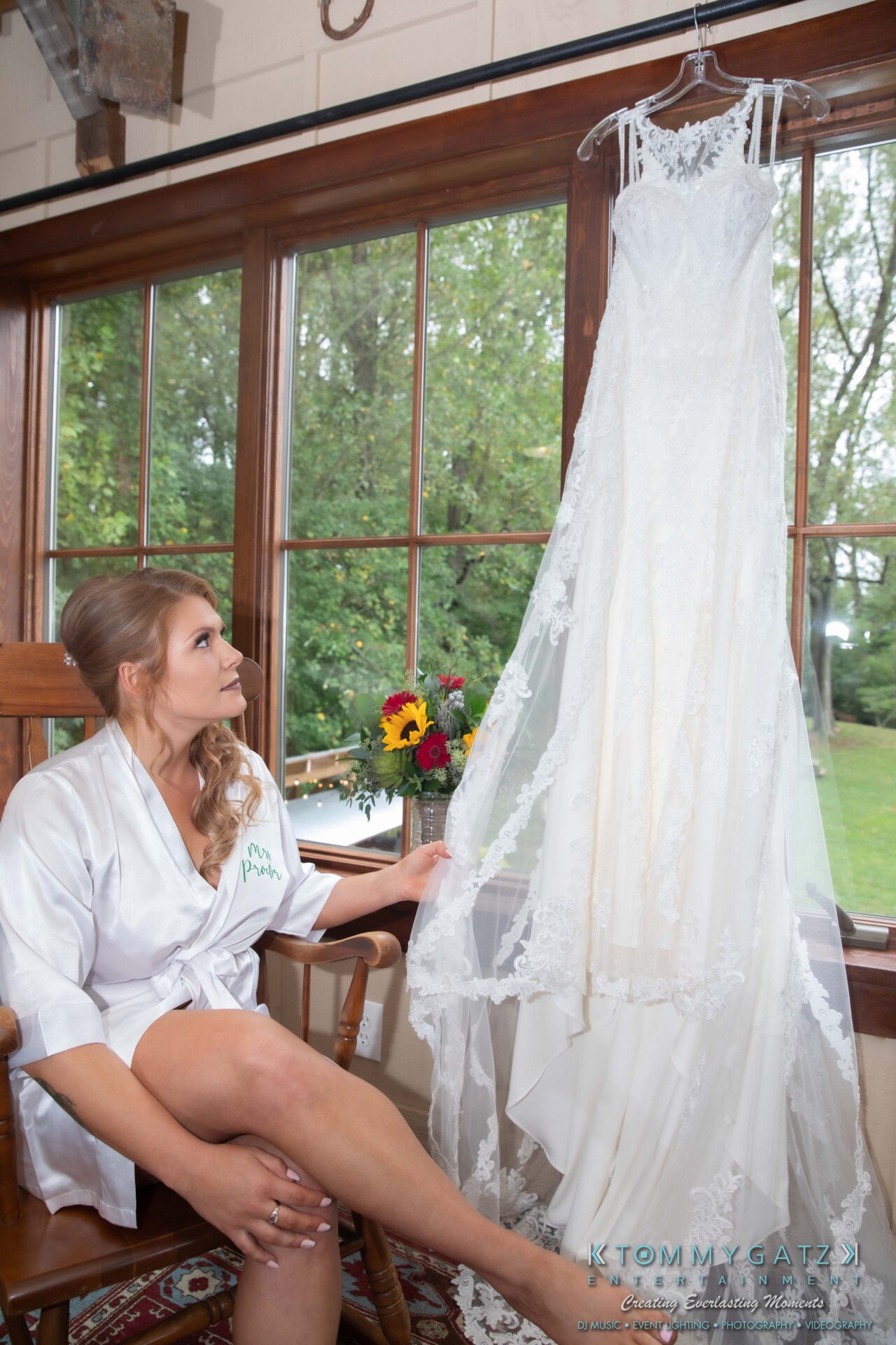 Proctor-Wedding-HIGHLIGHTS-091419-7 - TOMMY GATZ ENTERTAINMENT A woman in a white robe is seated and looking at a lace wedding dress hanging by a window.