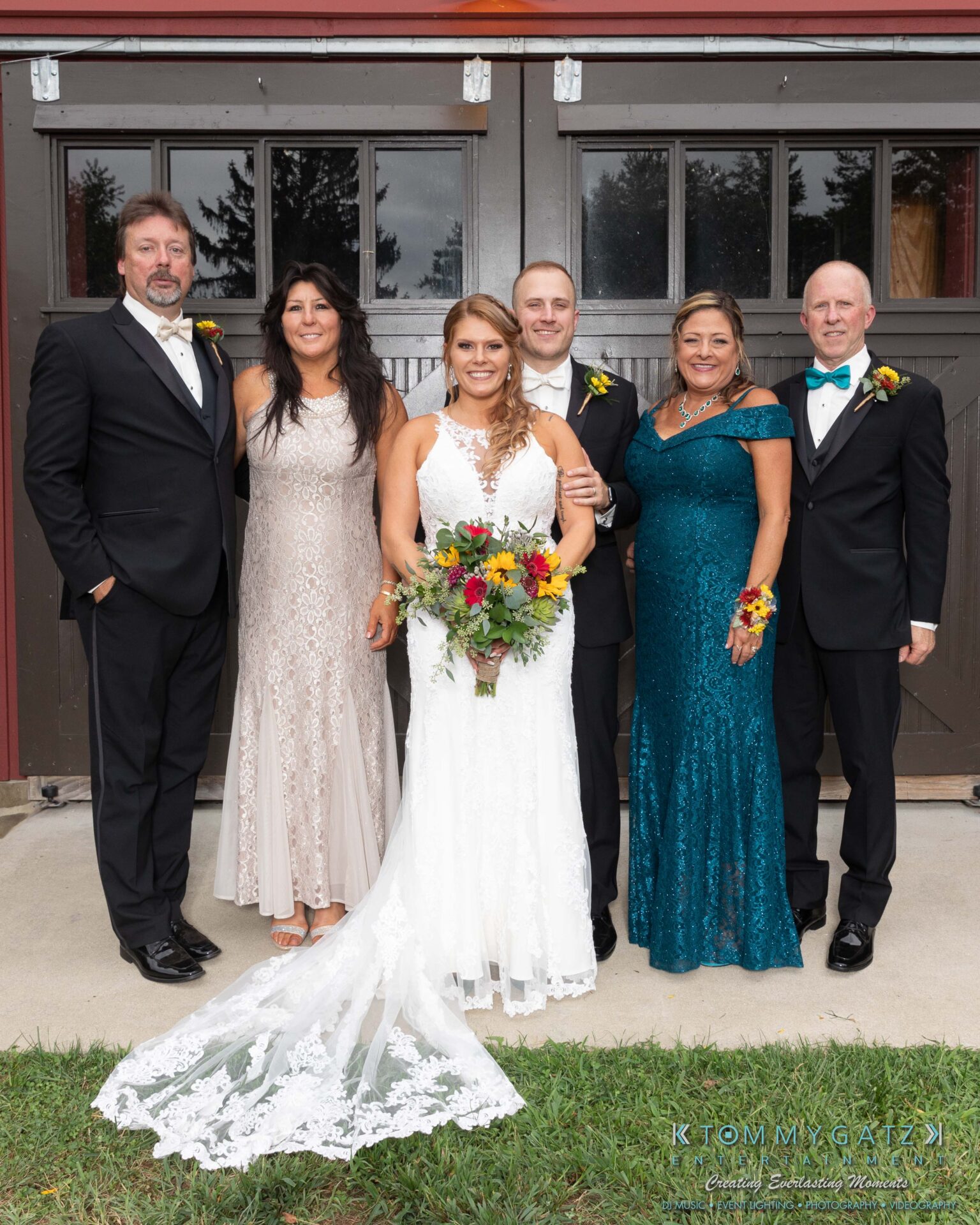 Proctor-Wedding-HIGHLIGHTS-091419-70 - TOMMY GATZ ENTERTAINMENT A bride and groom stand with four adults dressed formally, posing in front of a building with large doors. The bride holds a bouquet.