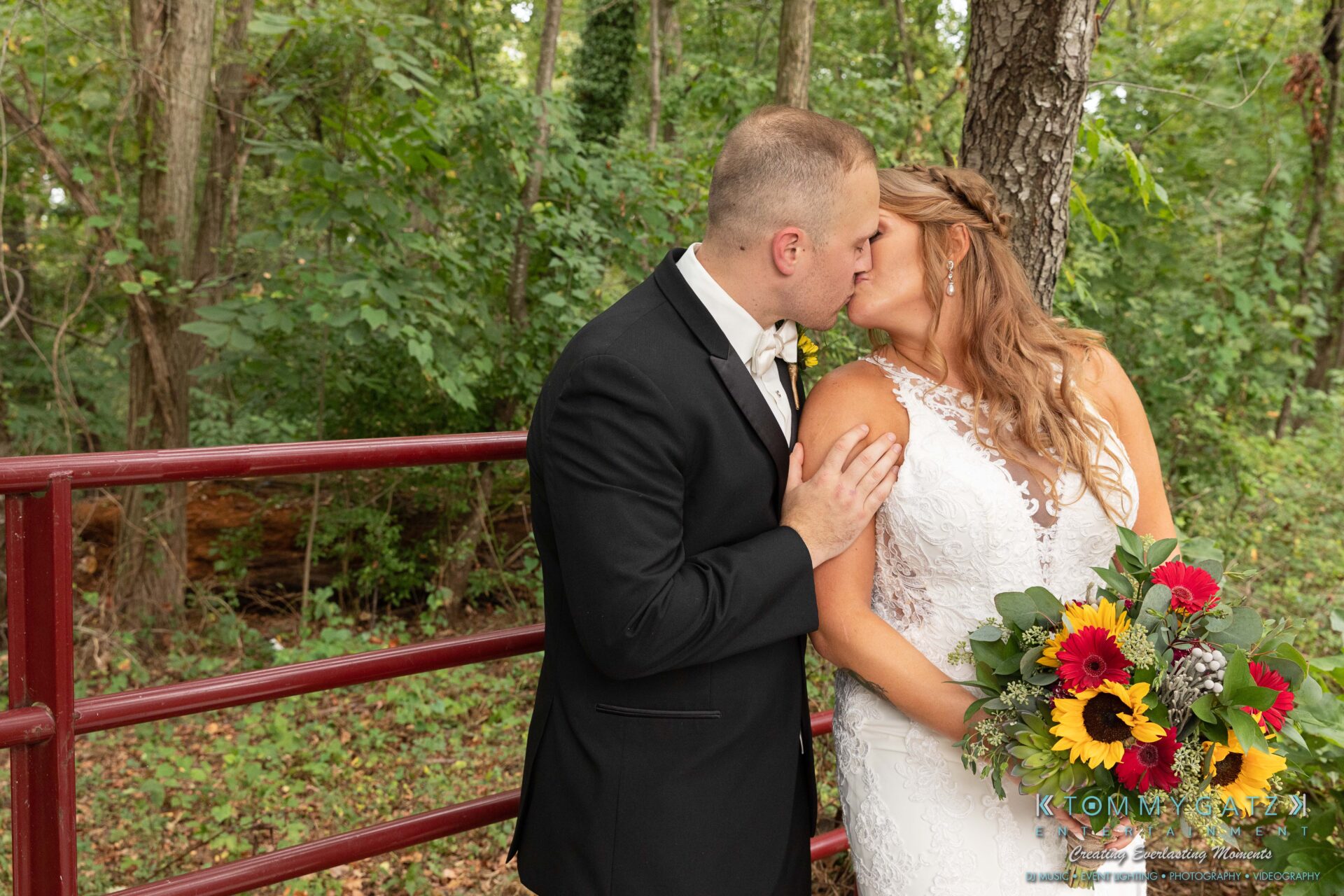 Proctor-Wedding-HIGHLIGHTS-091419-79 - TOMMY GATZ ENTERTAINMENT Bride and groom kiss outdoors in front of a red fence, surrounded by greenery. The bride holds a bouquet of yellow and red flowers.