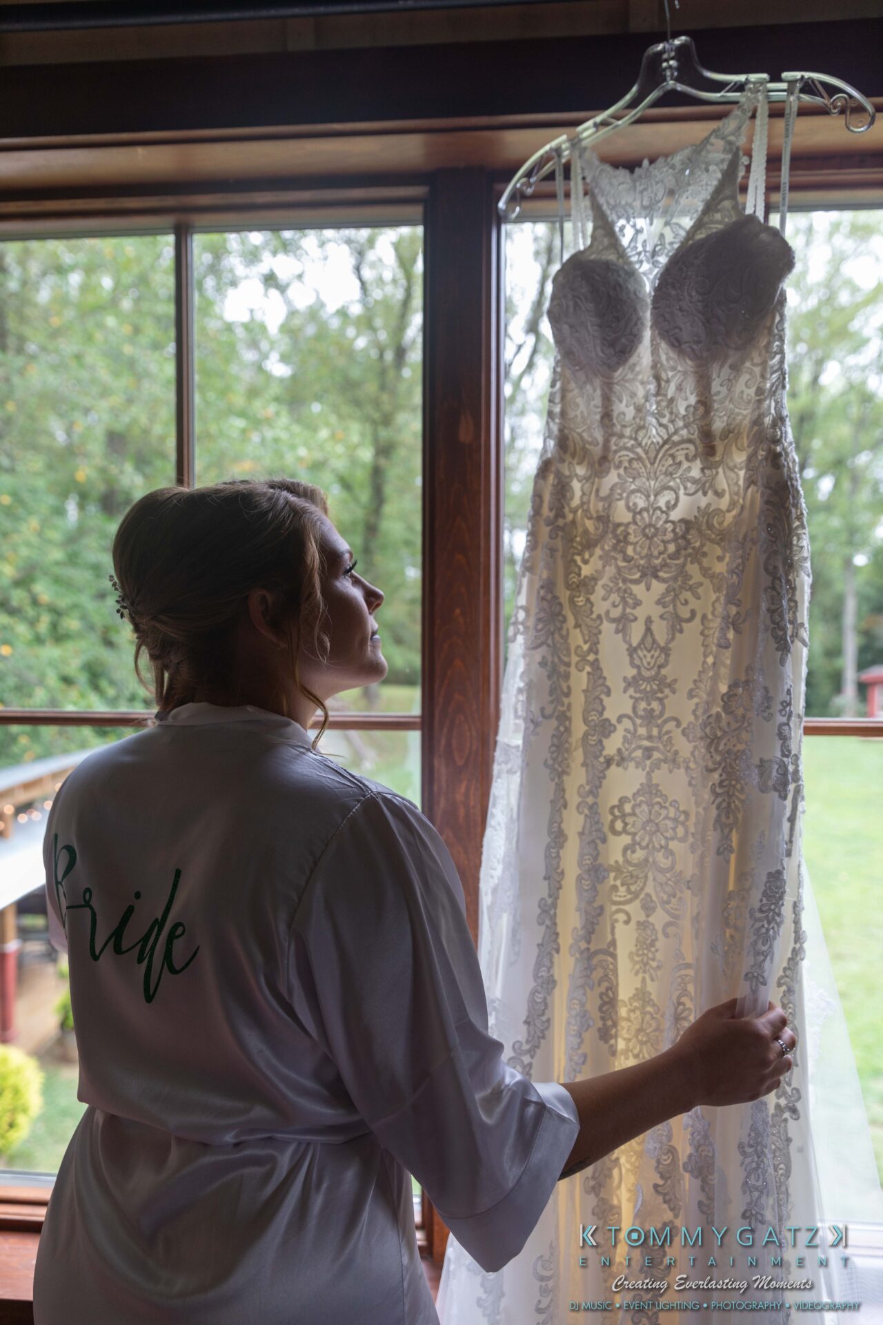 Proctor-Wedding-HIGHLIGHTS-091419-8 - TOMMY GATZ ENTERTAINMENT A woman in a "Bride" robe admires a lace wedding dress hanging by a window, with green foliage visible outside.