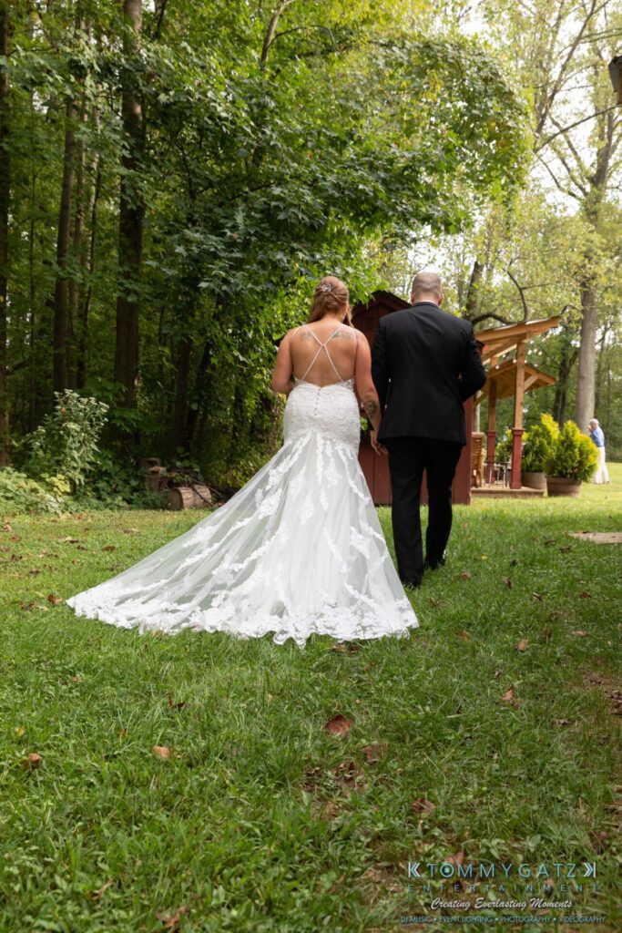Proctor-Wedding-HIGHLIGHTS-091419-81 - TOMMY GATZ ENTERTAINMENT A bride and groom walk hand in hand on a grassy path surrounded by trees. The bride wears a white dress with a long train, and the groom is in a black suit.