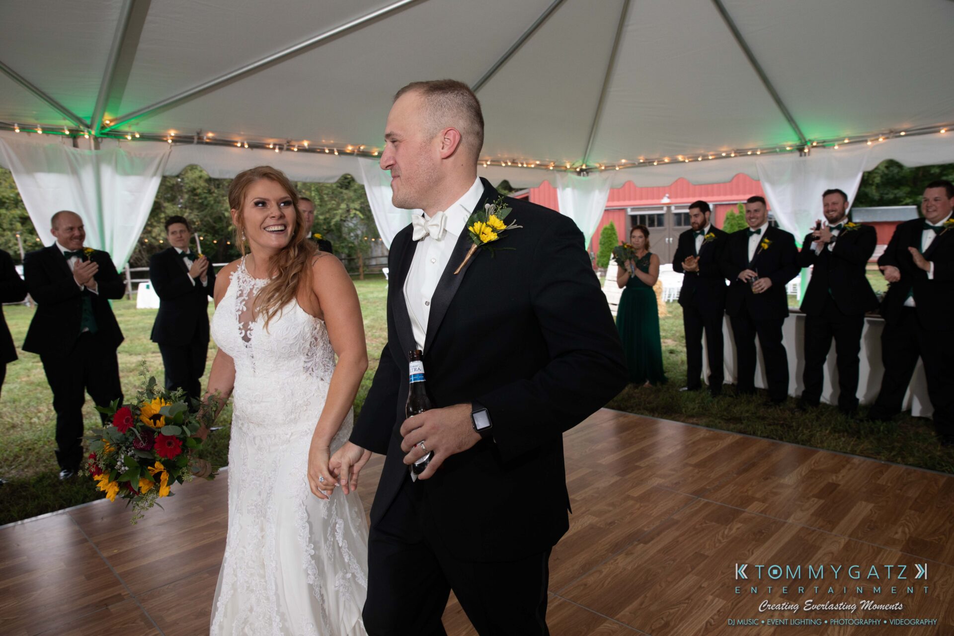 Proctor-Wedding-HIGHLIGHTS-091419-83 - TOMMY GATZ ENTERTAINMENT Bride and groom walk hand in hand under a tent. The groom holds a beer bottle, and the bridal party applauds in the background.