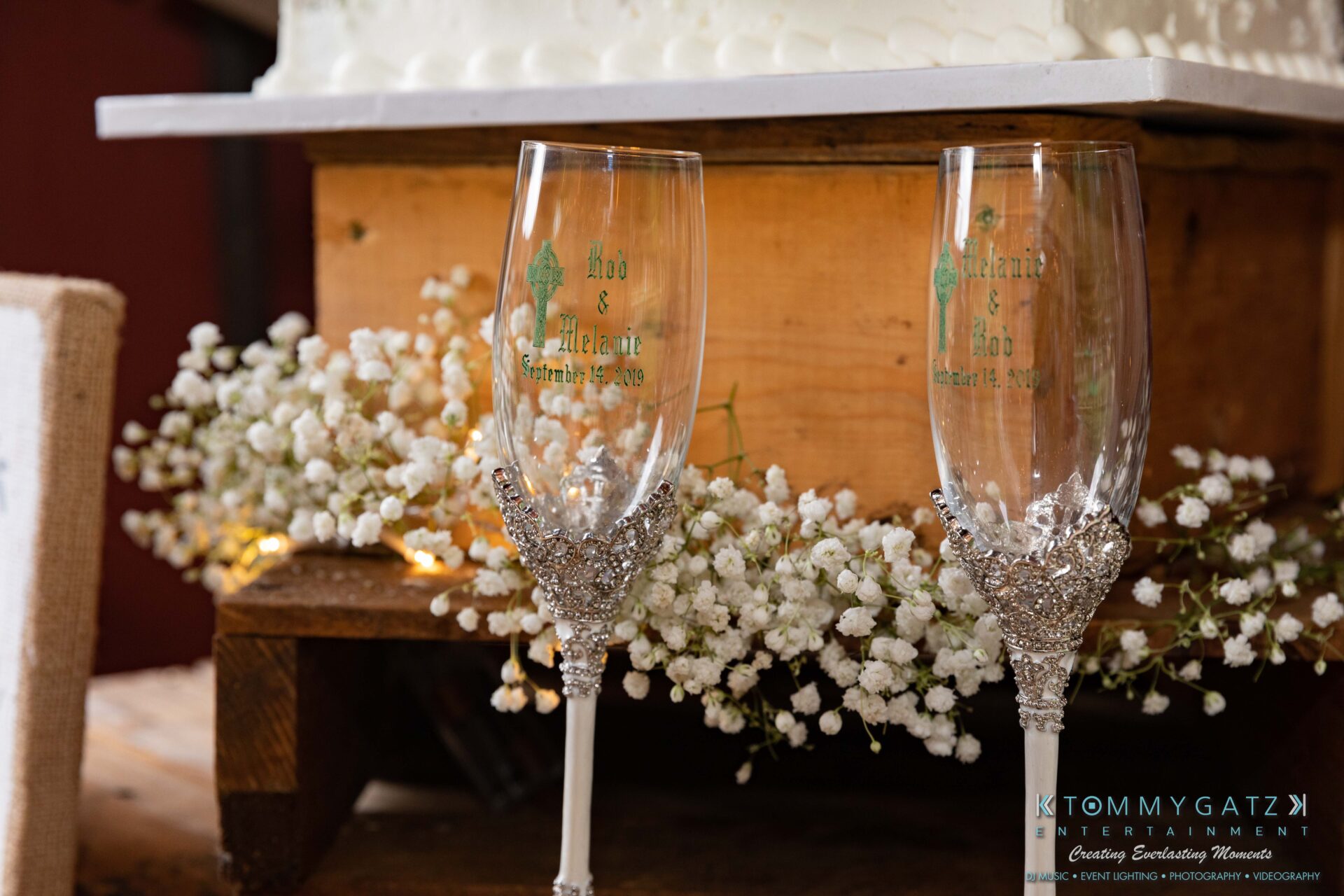 Proctor-Wedding-HIGHLIGHTS-091419-86 - TOMMY GATZ ENTERTAINMENT Two engraved champagne glasses with silver bases are placed in front of a wooden stand adorned with white flowers. A wedding cake is partially visible on the stand.