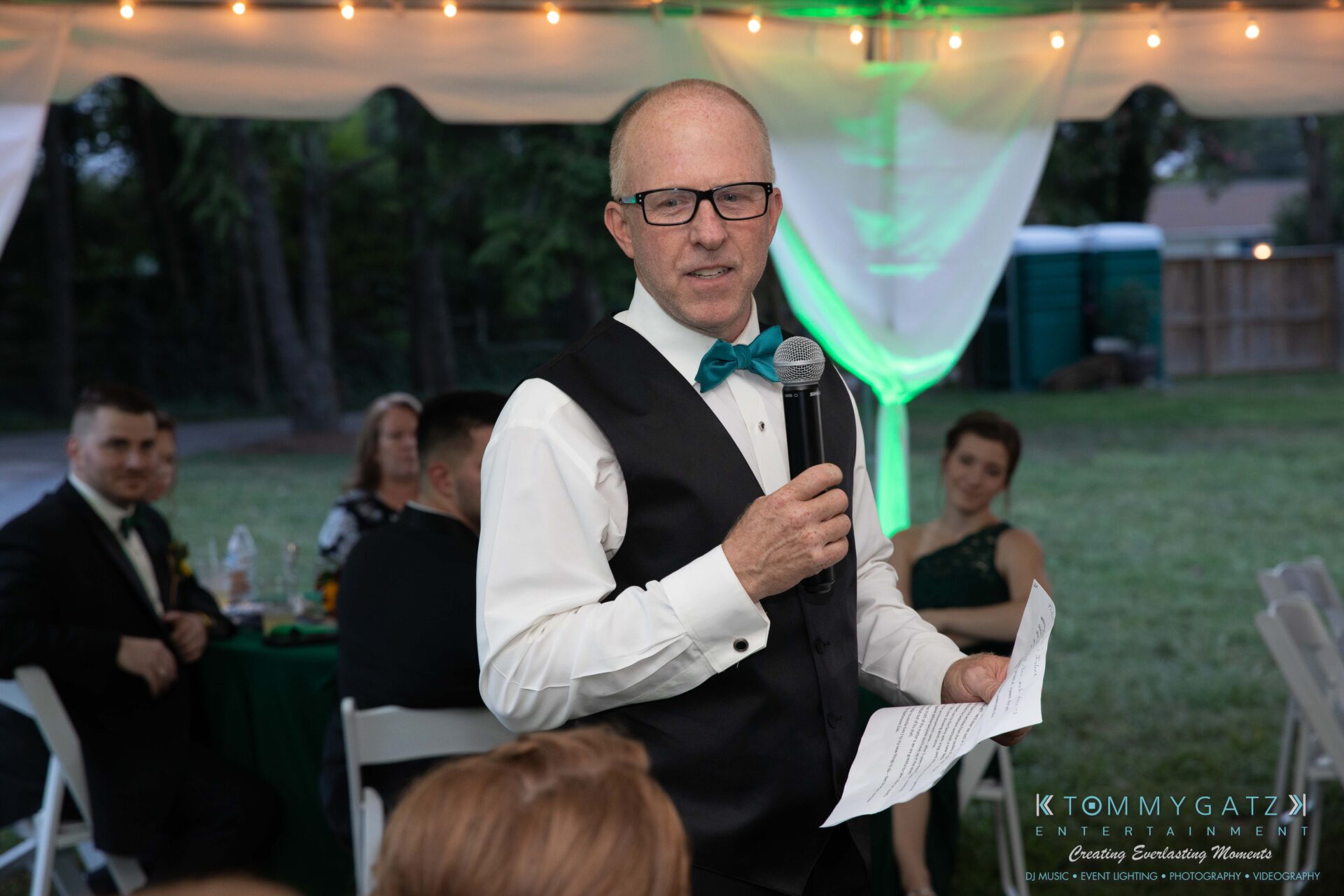 Proctor-Wedding-HIGHLIGHTS-091419-87 - TOMMY GATZ ENTERTAINMENT A man in formal attire speaks into a microphone while holding a piece of paper at an outdoor event. The background includes seated guests and a tent.