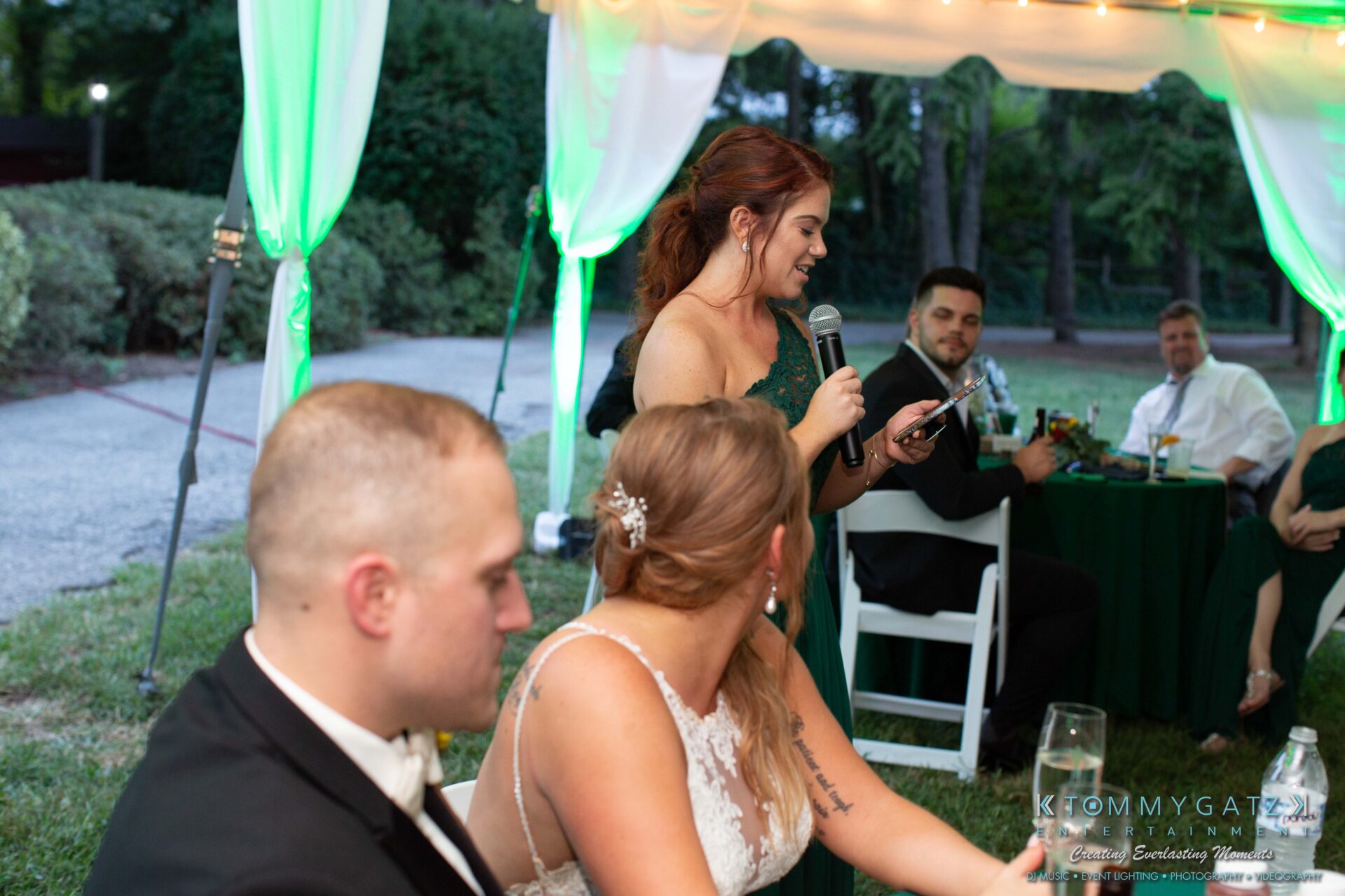Proctor-Wedding-HIGHLIGHTS-091419-89 - TOMMY GATZ ENTERTAINMENT A woman in a green dress speaks into a microphone at an outdoor event. Seated guests listen at tables with green tablecloths.